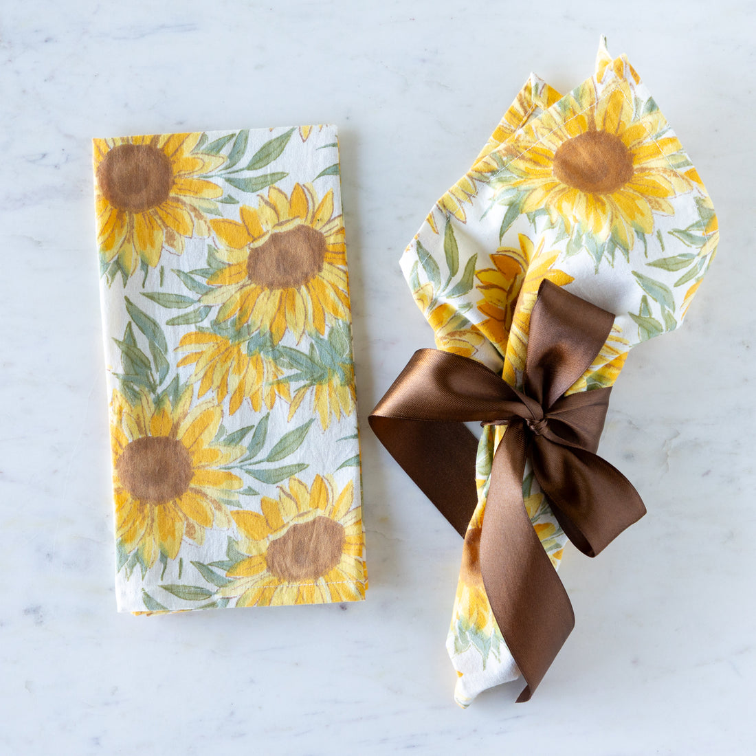 Sunflower-patterned napkins, one tied with a brown ribbon, the other folded on a marble table.