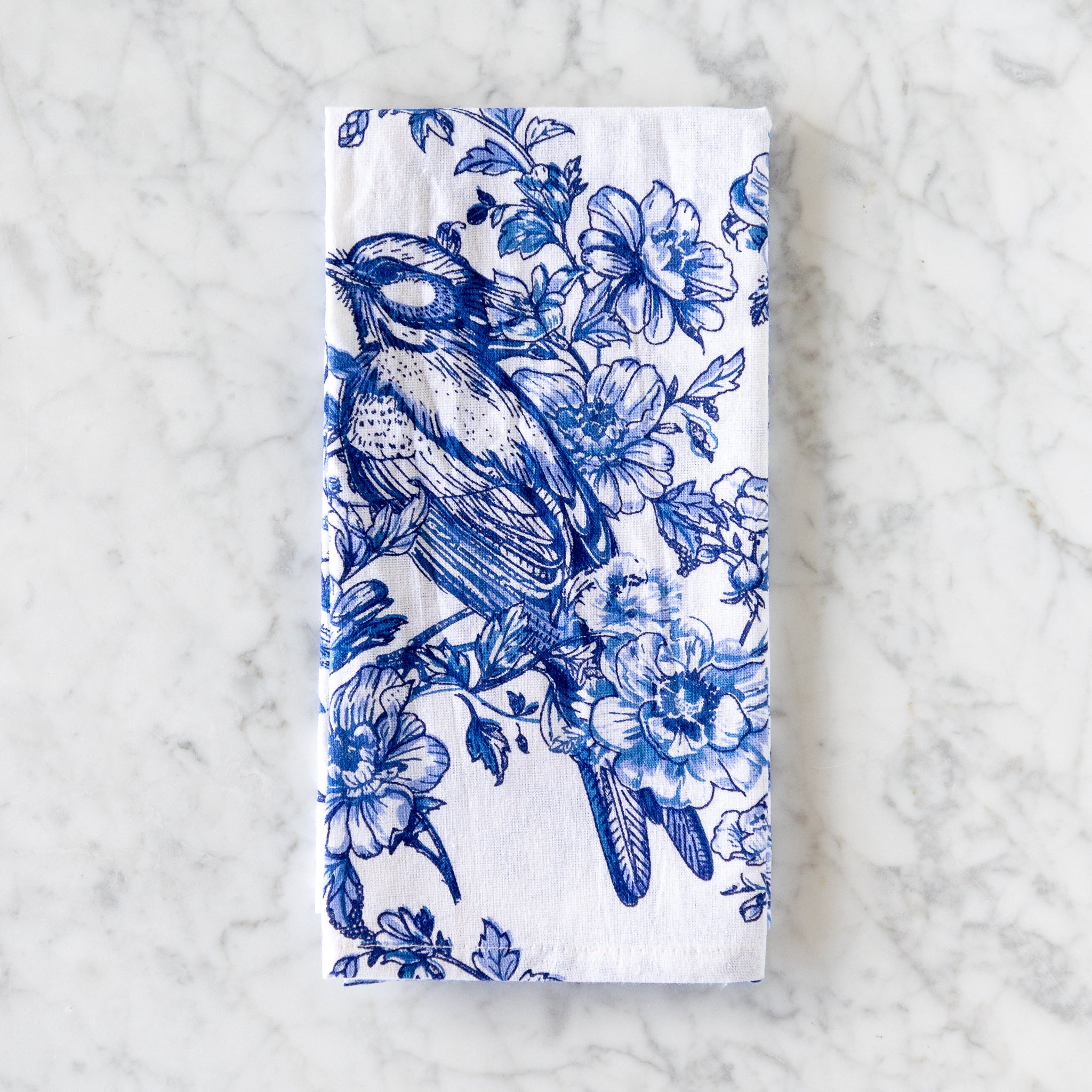 A white cotton napkin, made in India, features a blue illustration of a bird perched among flowers and is displayed on a marble table.