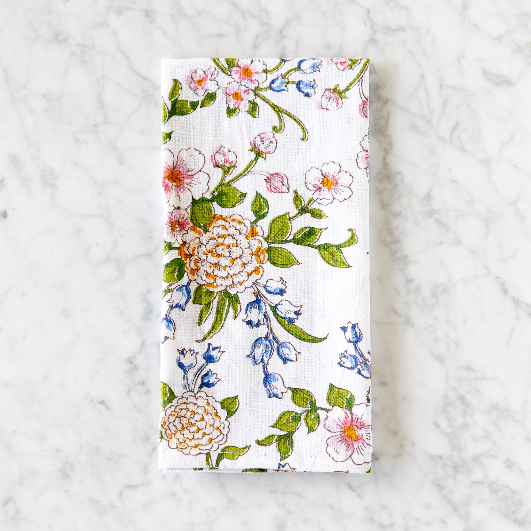 A white cotton napkin, made in India, features a colorful illustration of flowers and leaves and is displayed on a marble table.