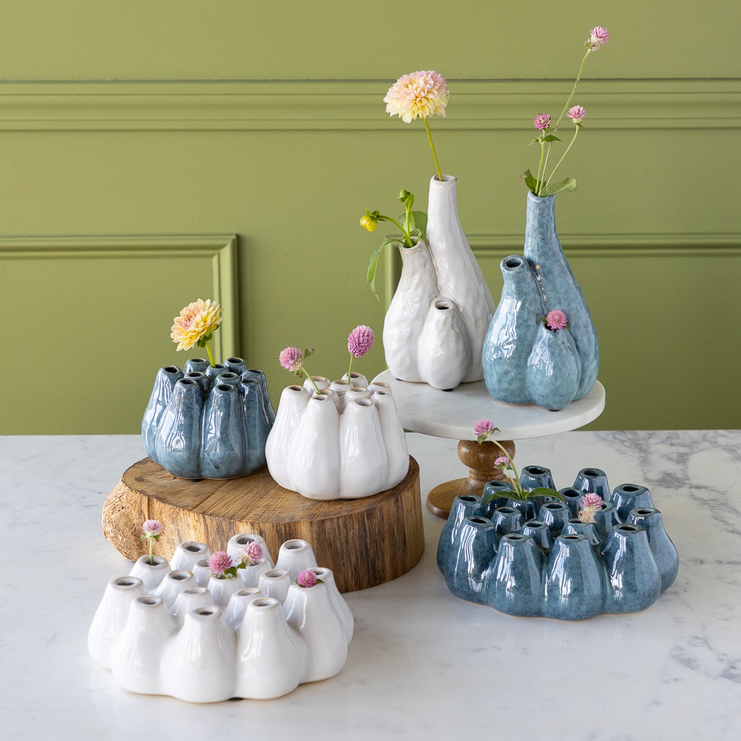 Collection of ceramic vases with flowers on a marble surface against a green wall.
