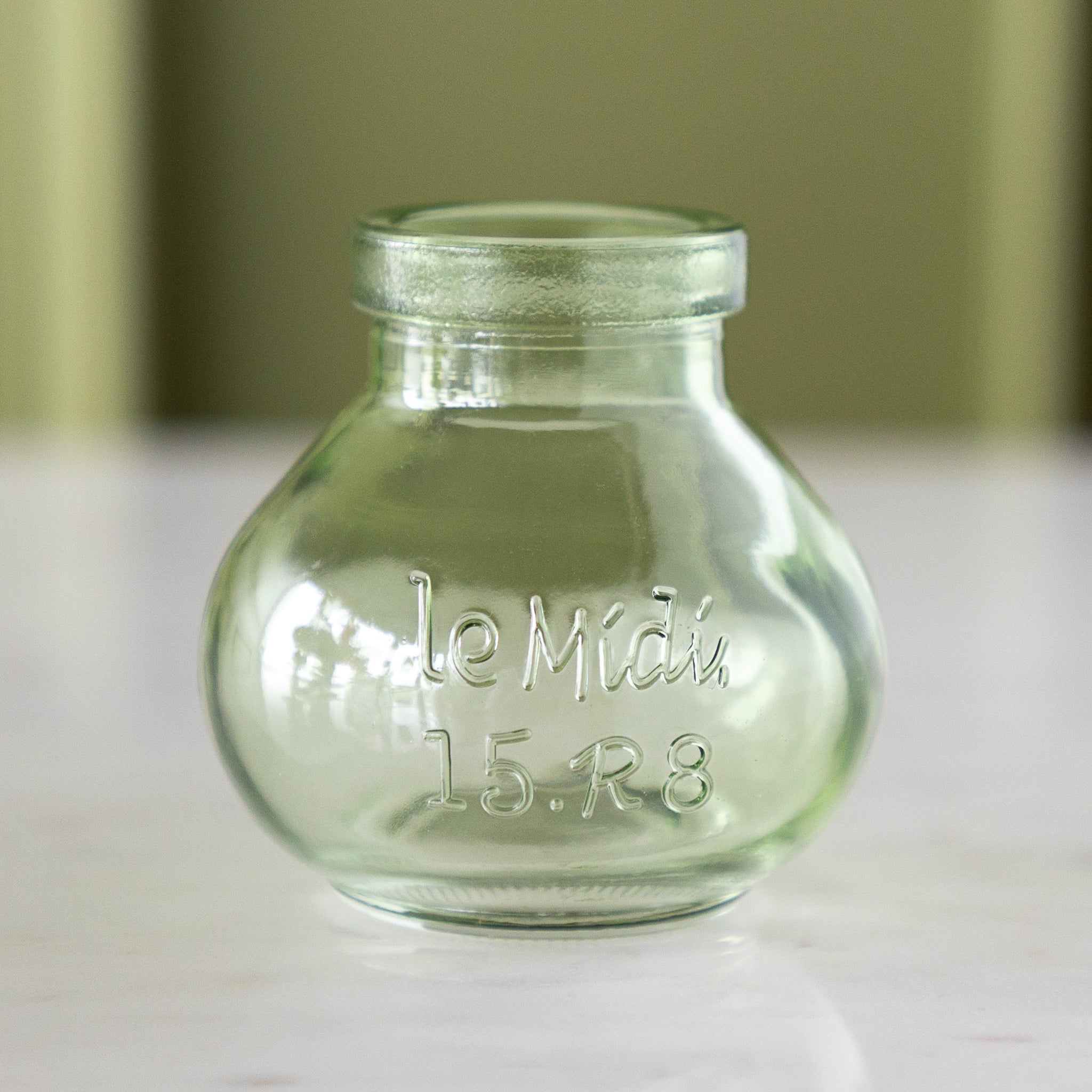 A small, empty, clear green glass bottle with "le Midi. 15.R8" embossed on the front, reminiscent of vintage bud vases, stands on a marble table against a light green background.