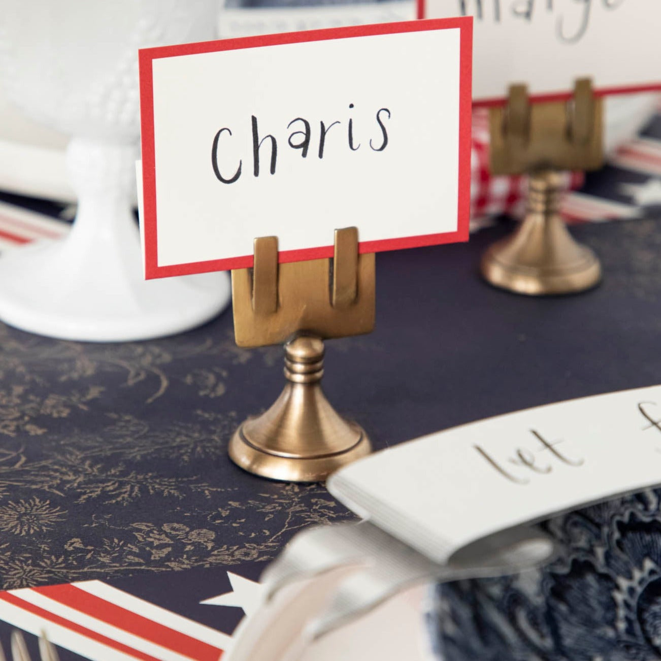 Close-up of a Red Frame Place Card labeled "Charis" held by a brass table card holder on a nautical-themed table setting.