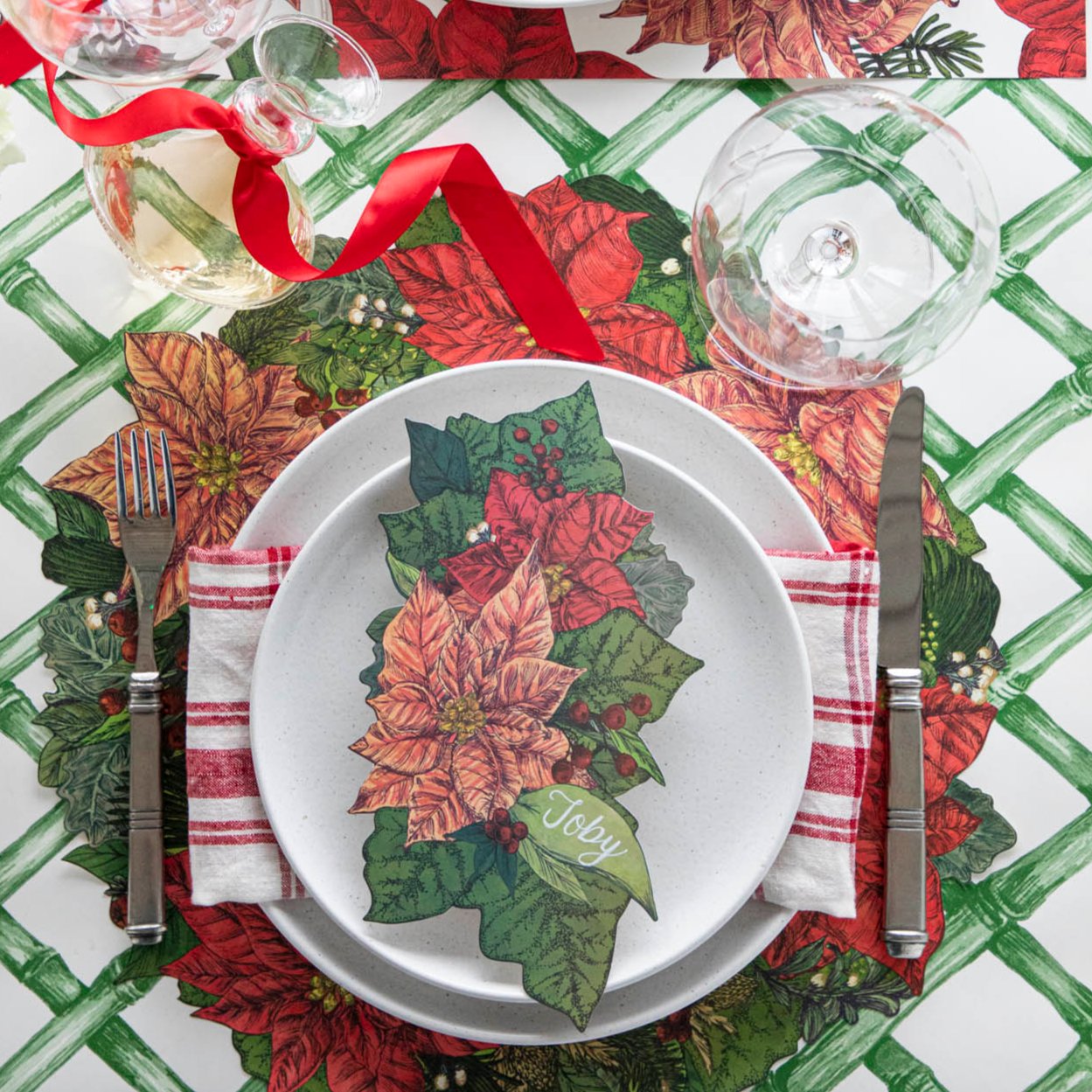 An elegant table setting with the Die-cut Poinsettia Placemat on top of the Green Lattice Runner, with the Poinsettia Table Accent with the name "Toby" written on it, on top of a plate.