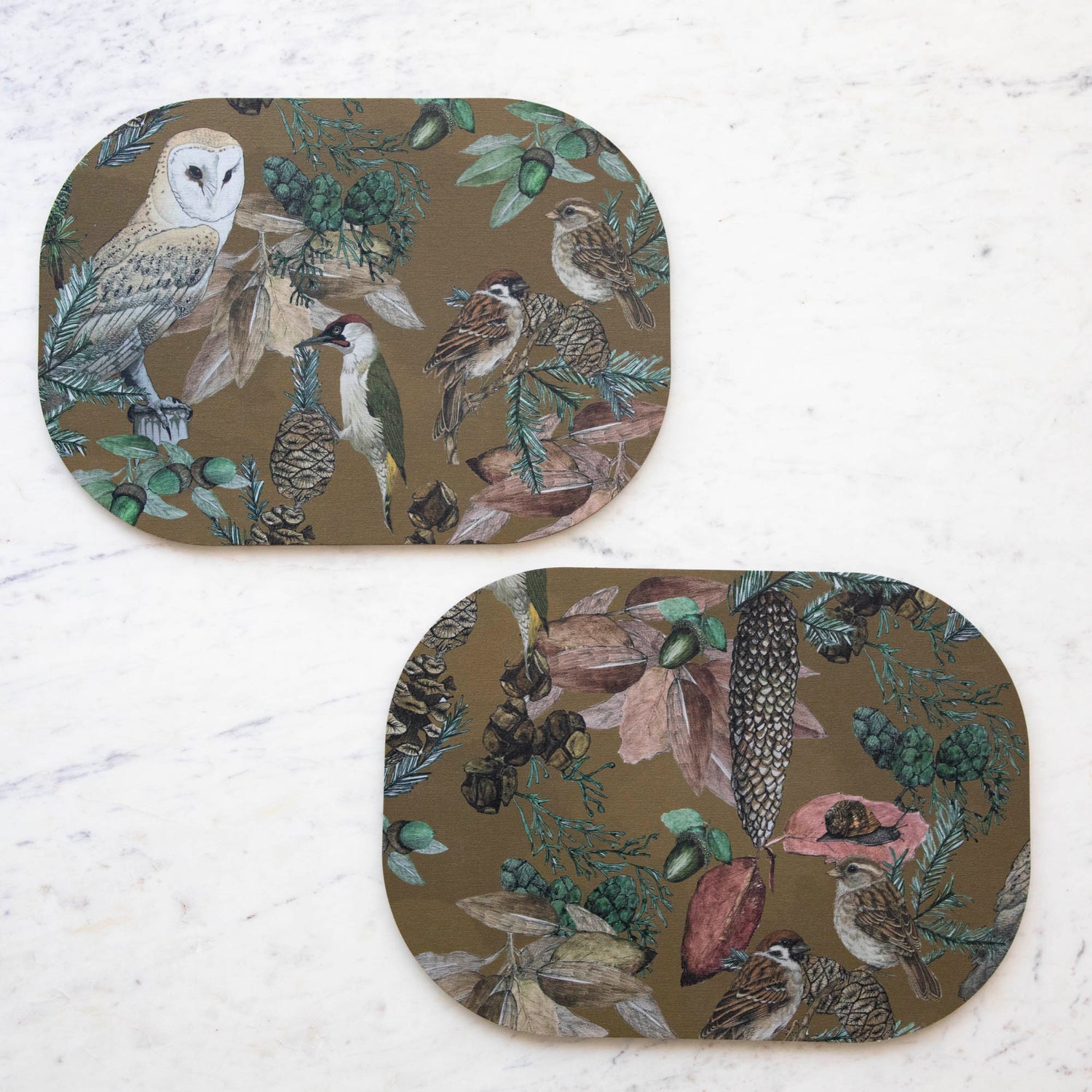 Two oval placemats with a bird, floral, and butterfly pattern, displayed on a table with striped tablecloth.