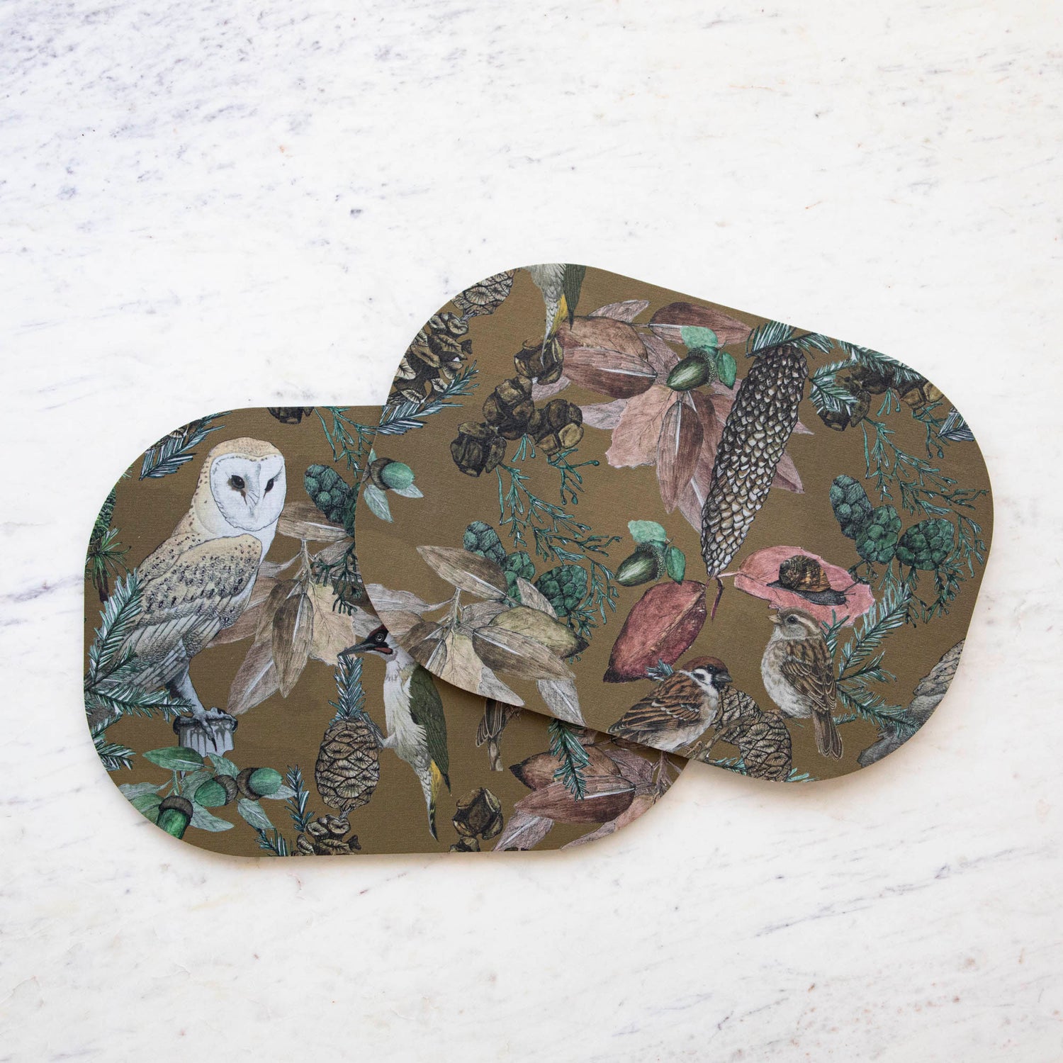 Two oval placemats with a bird, floral, and butterfly pattern, displayed on a table with striped tablecloth.