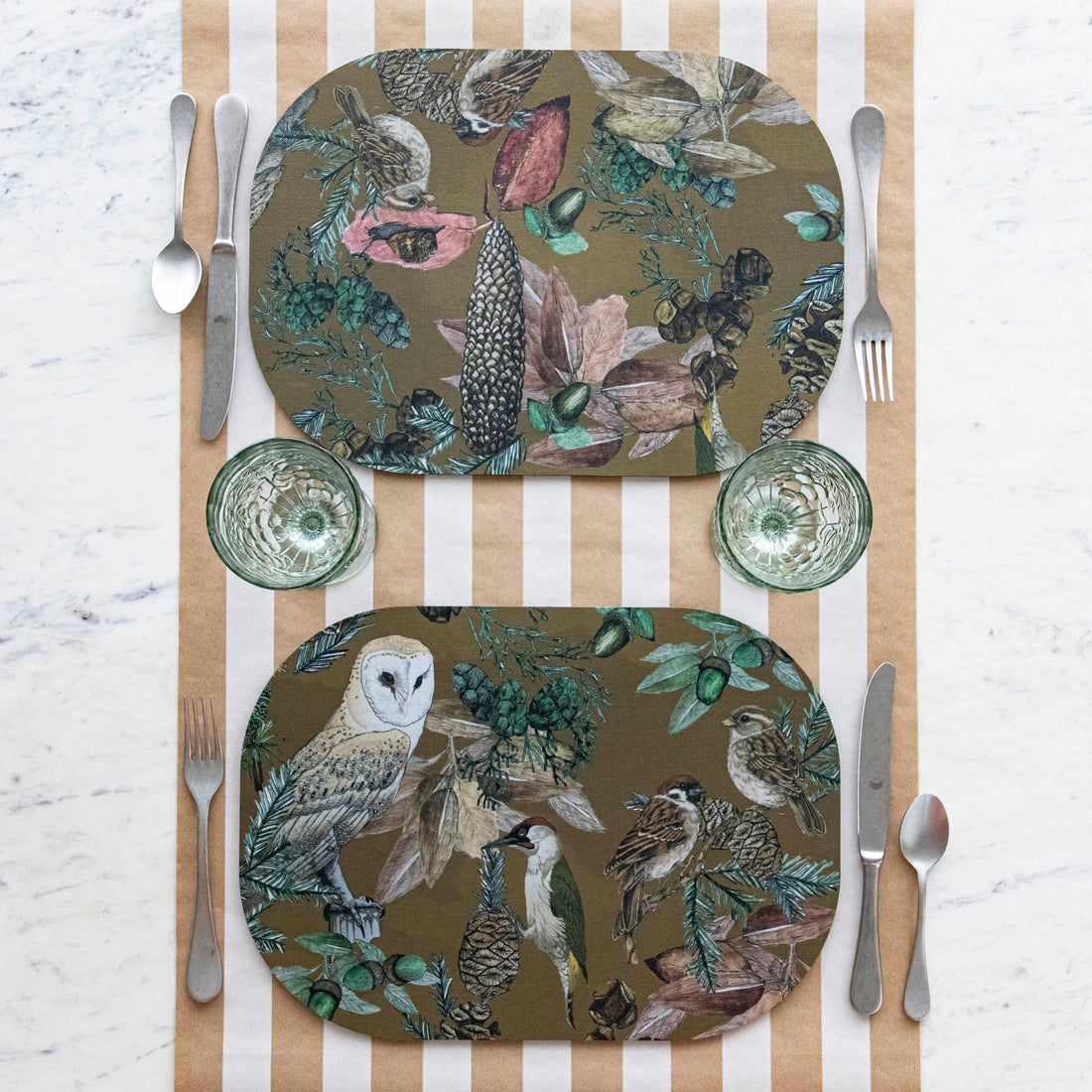 Two oval placemats with a bird, floral, and butterfly pattern, displayed on a table with striped tablecloth.