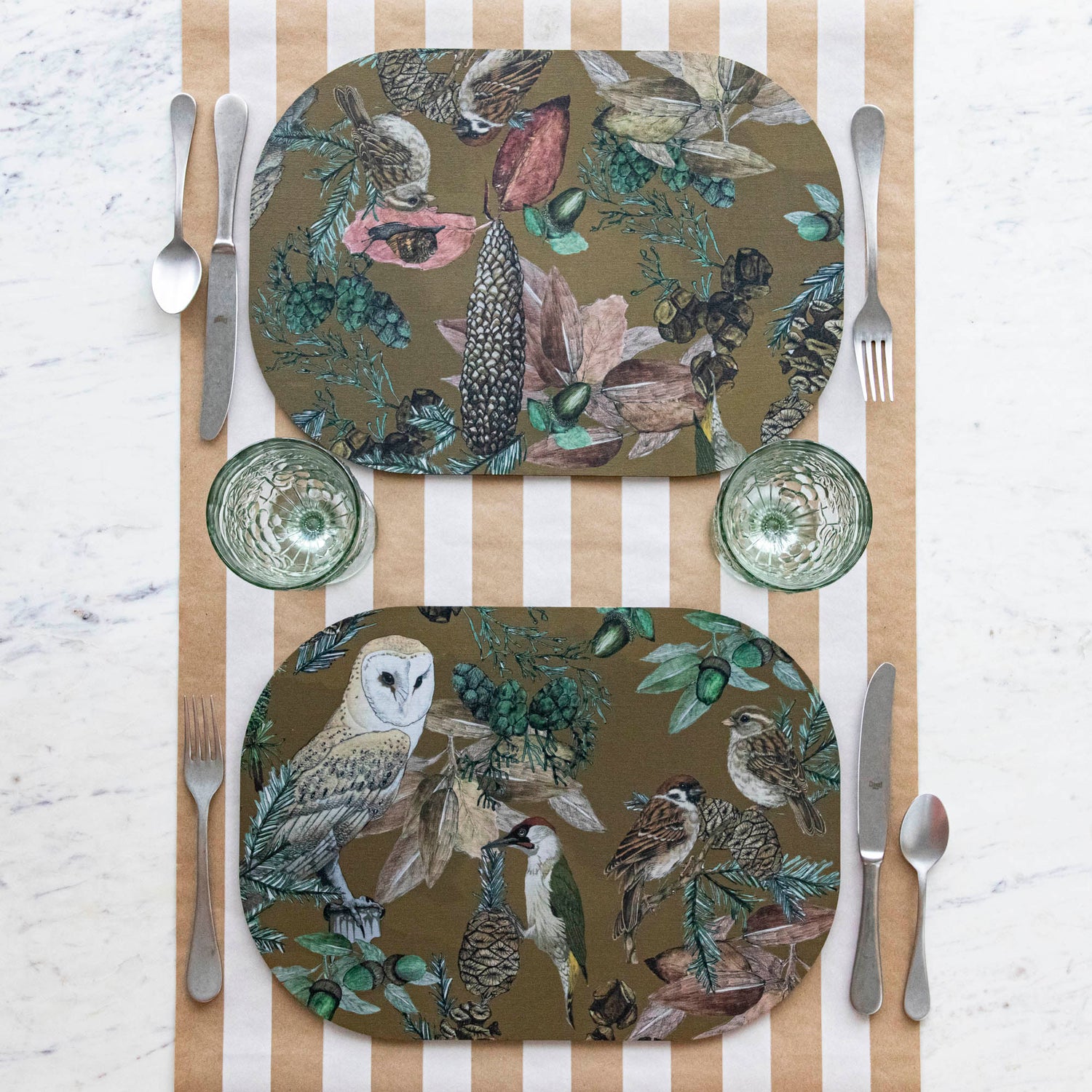 Two oval placemats with a bird, floral, and butterfly pattern, displayed on a table with striped tablecloth.