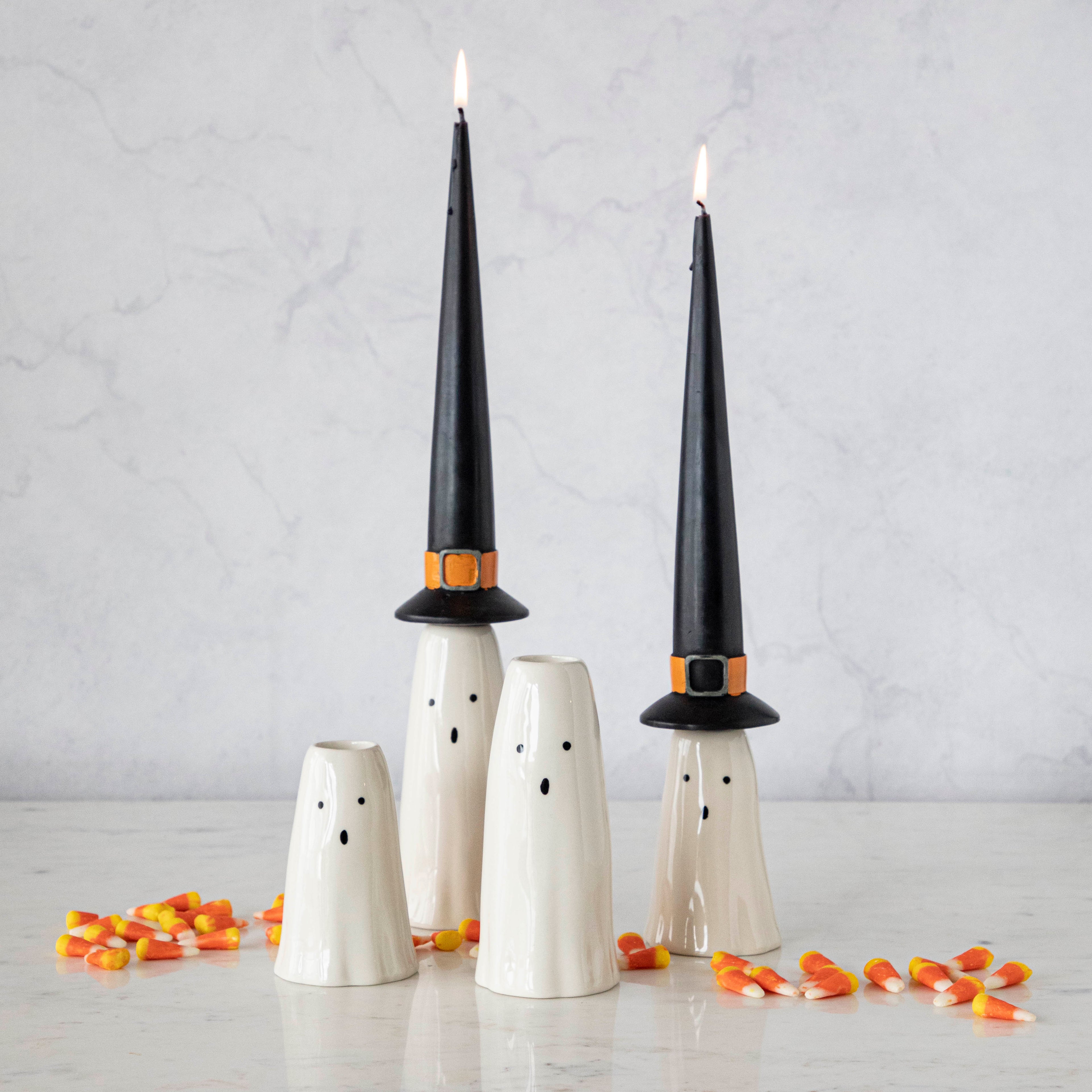 Four ghost candle holders, two of which hold lit witch hat taper candles. 
