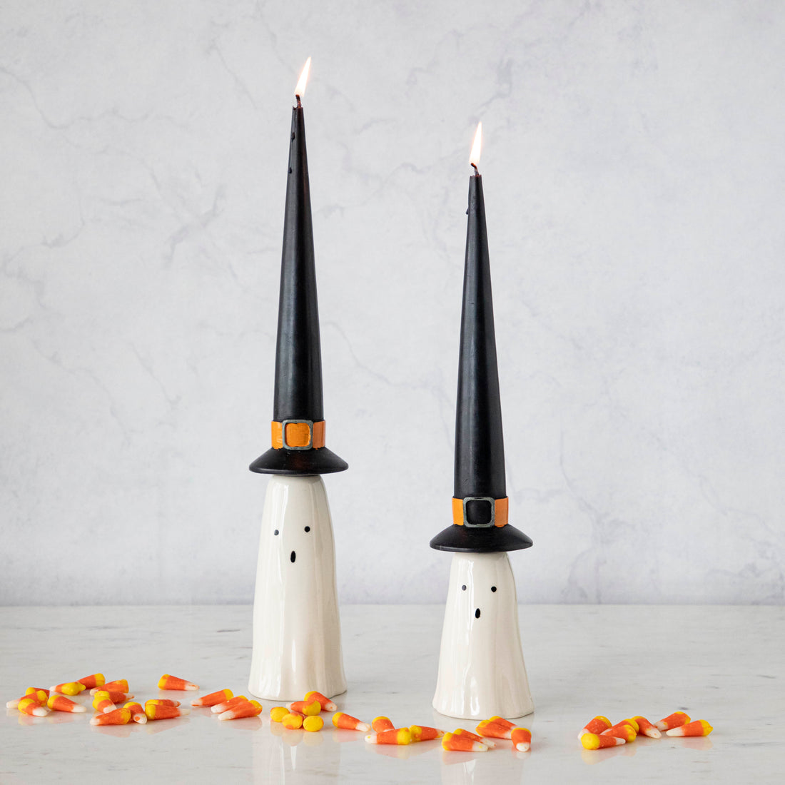 Two taper candles designed to look like ghosts with black witch hats, accompanied by scattered candy corn on a light surface.