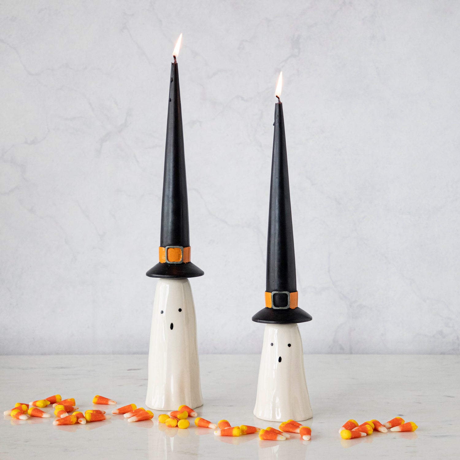 Two taper candles designed to look like ghosts with black witch hats, accompanied by scattered candy corn on a light surface.