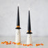 Two taper candles designed to look like ghosts with black witch hats, accompanied by scattered candy corn on a light surface.