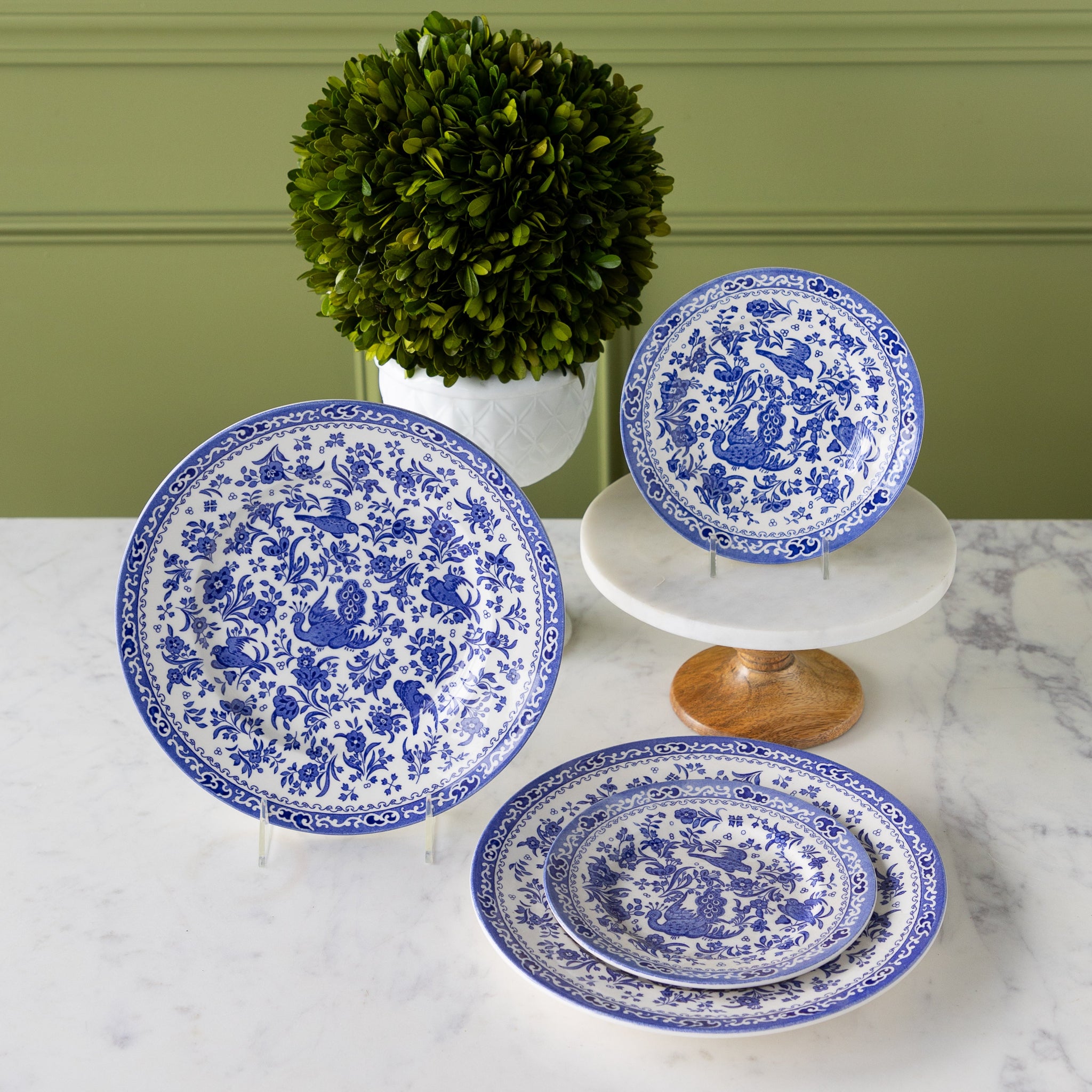 Burleigh Blue Regal Peacock Dinnerware propped on stands in front of a topiary plant.