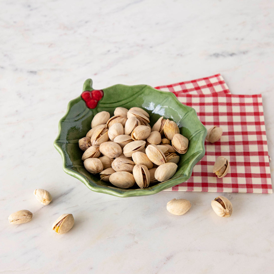 A green stoneware bowl with a holly leaf pattern filled with pistachios, placed on a red and white checkered napkin on a marble surface.