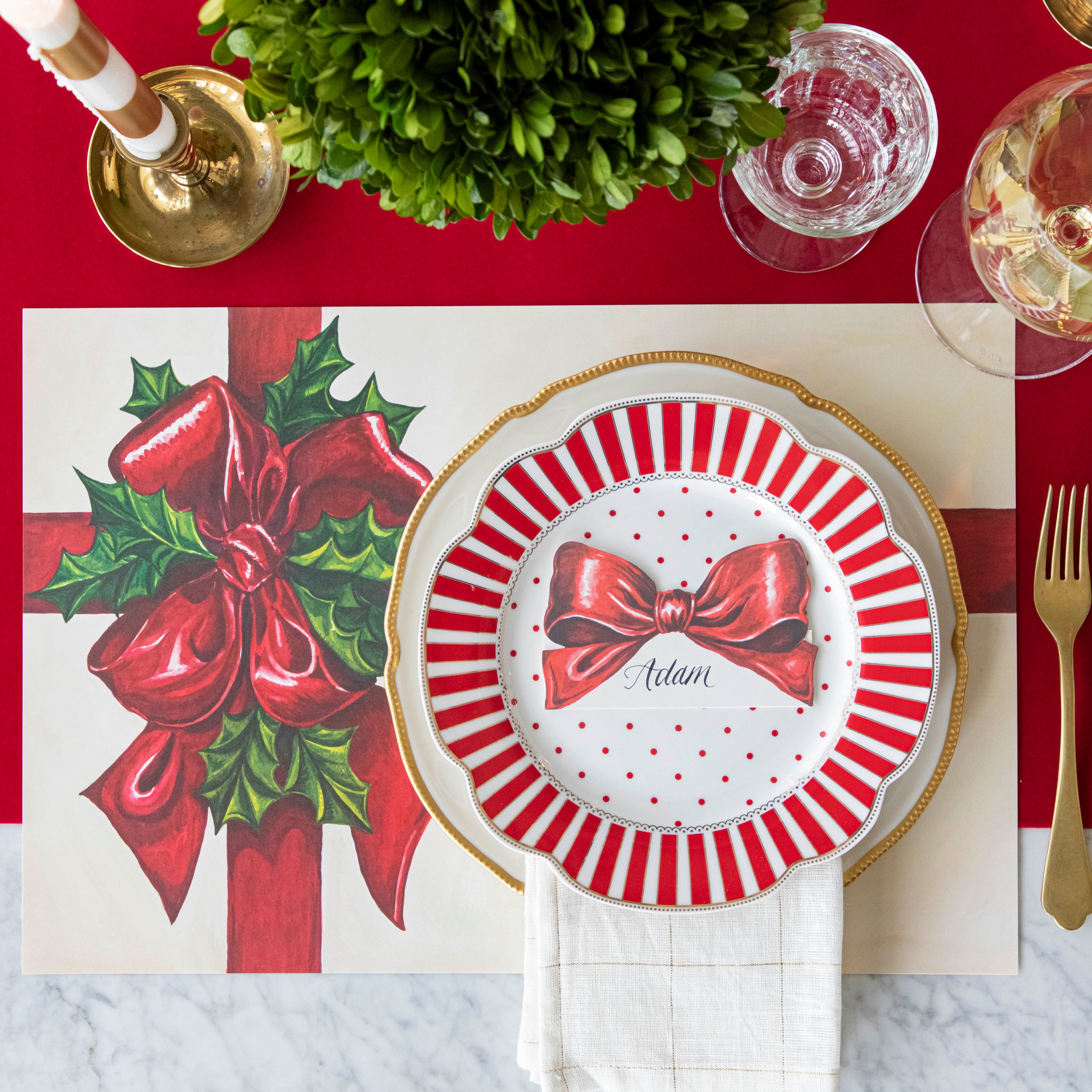 An elegant, Christms themed table setting with the Bow Place Card with "Adam" written on it in cursive, resting on two dinner plates, on top of the Christmas Present Placemat and Red Flocked Runner.