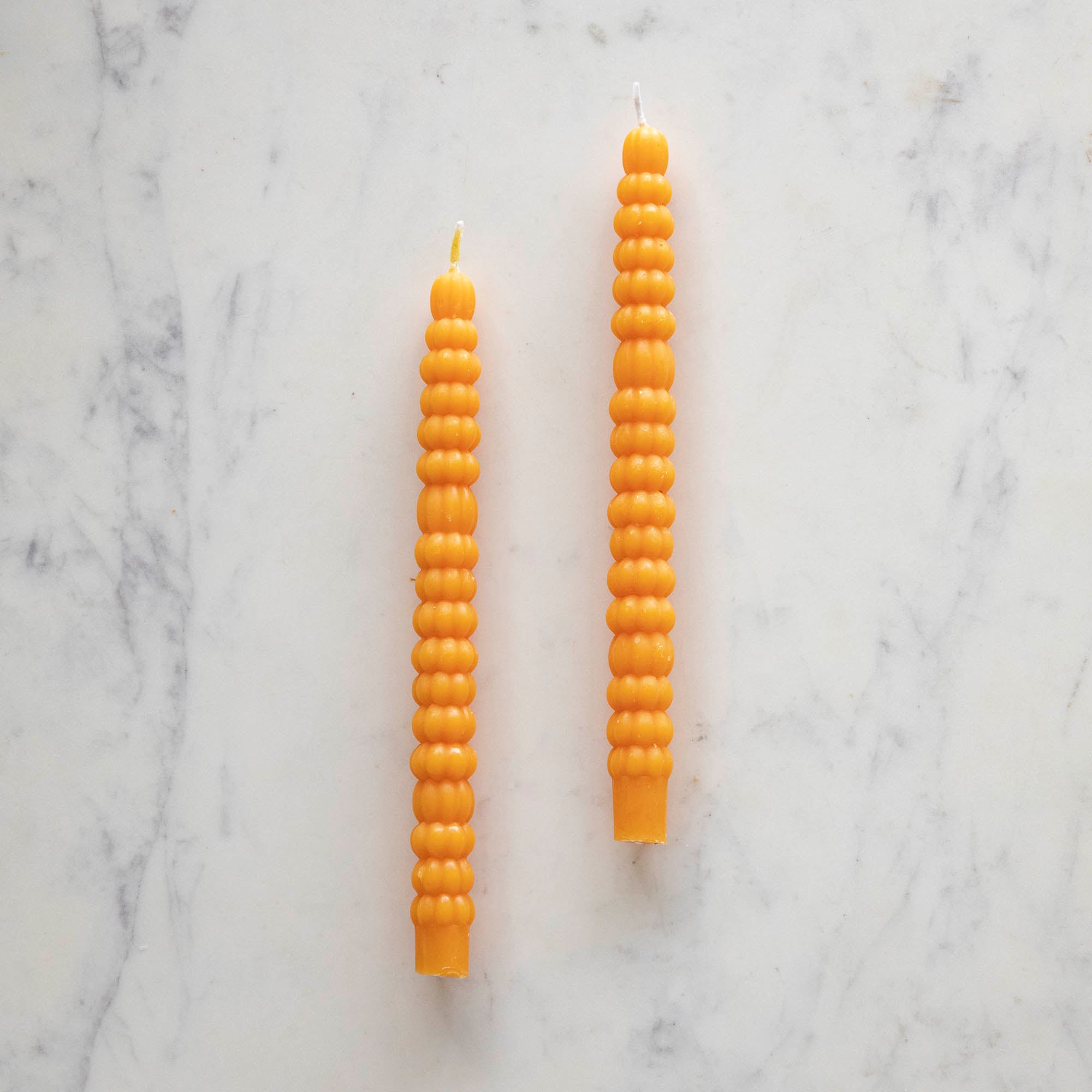 Two orange taper candles resembling stacks of tiny pumpkins laying on a white table.