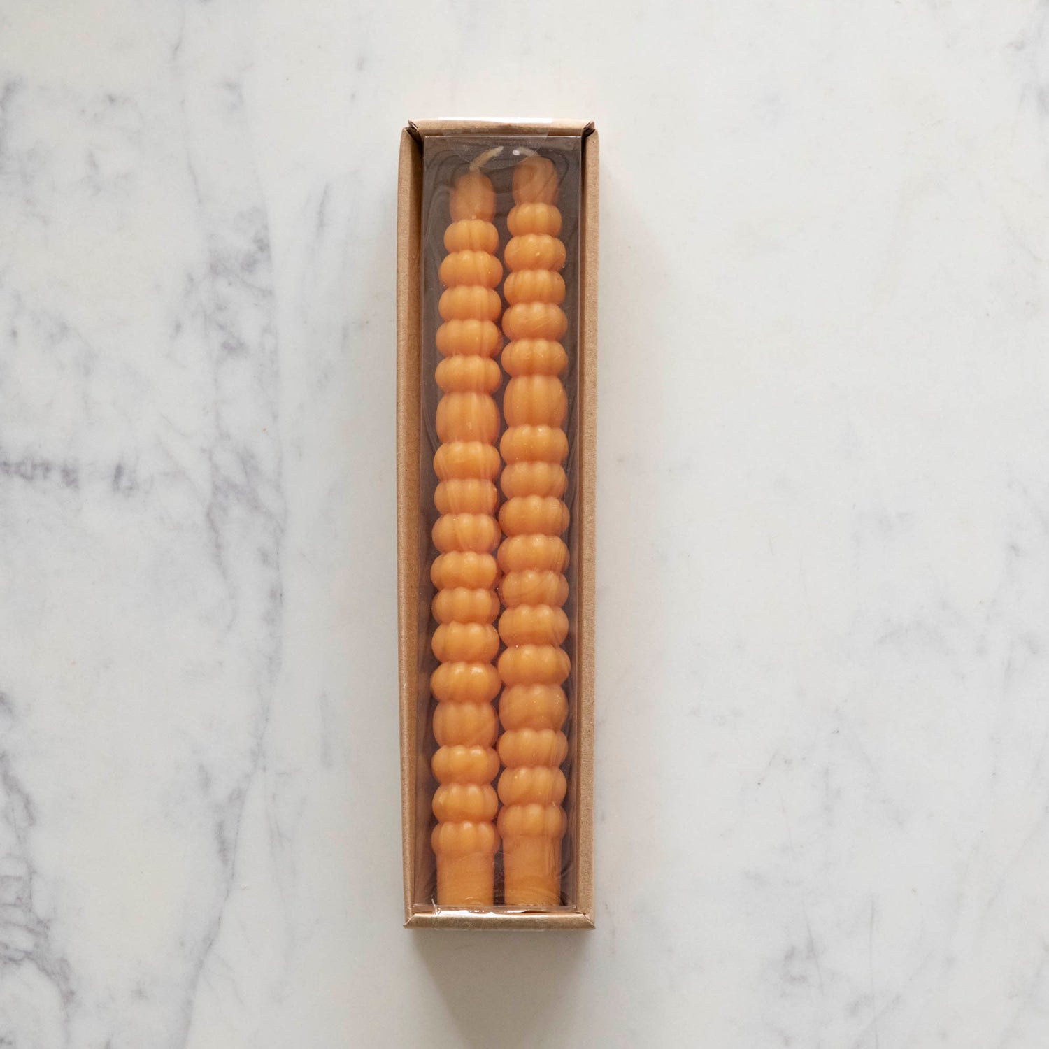 Two orange taper candles resembling stacks of tiny pumpkins laying in their package on a white table. 