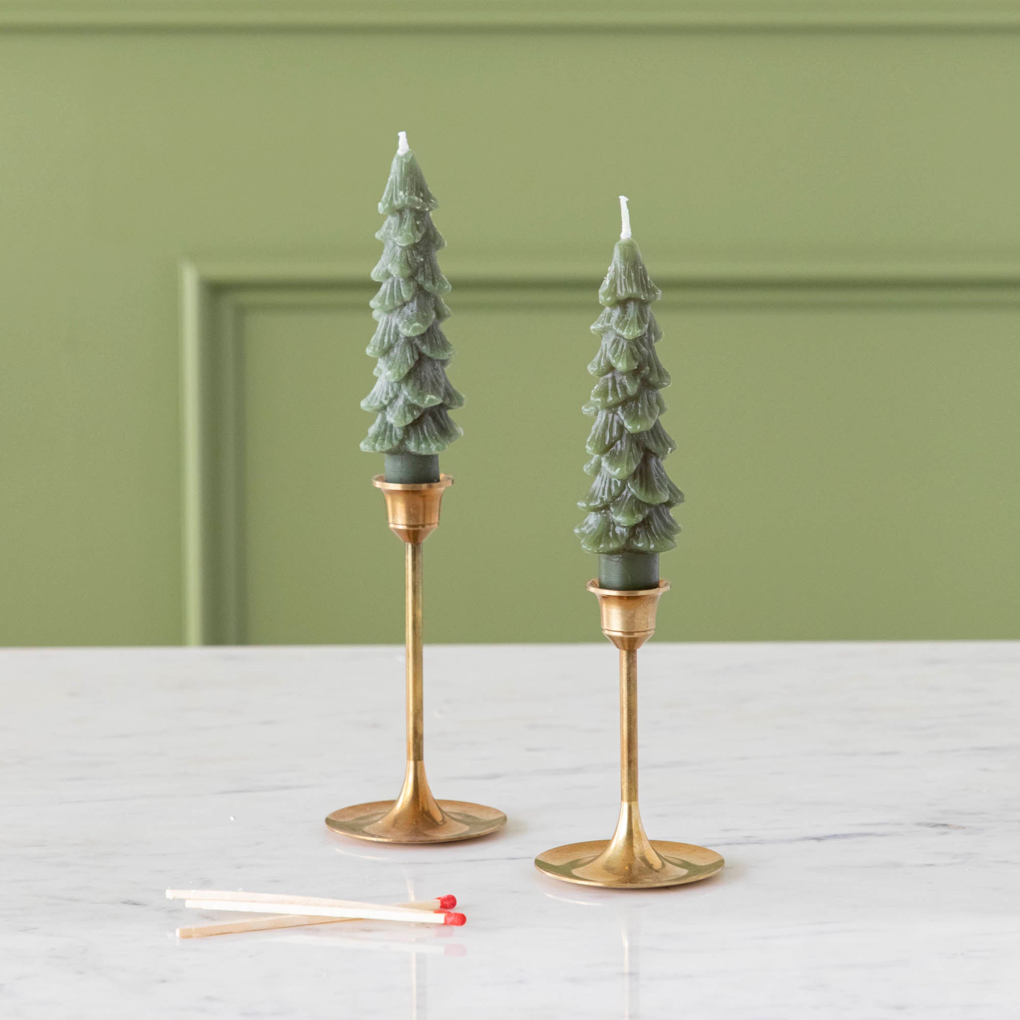 A set of two green tree-shaped taper candles placed on gold-colored candle holders.