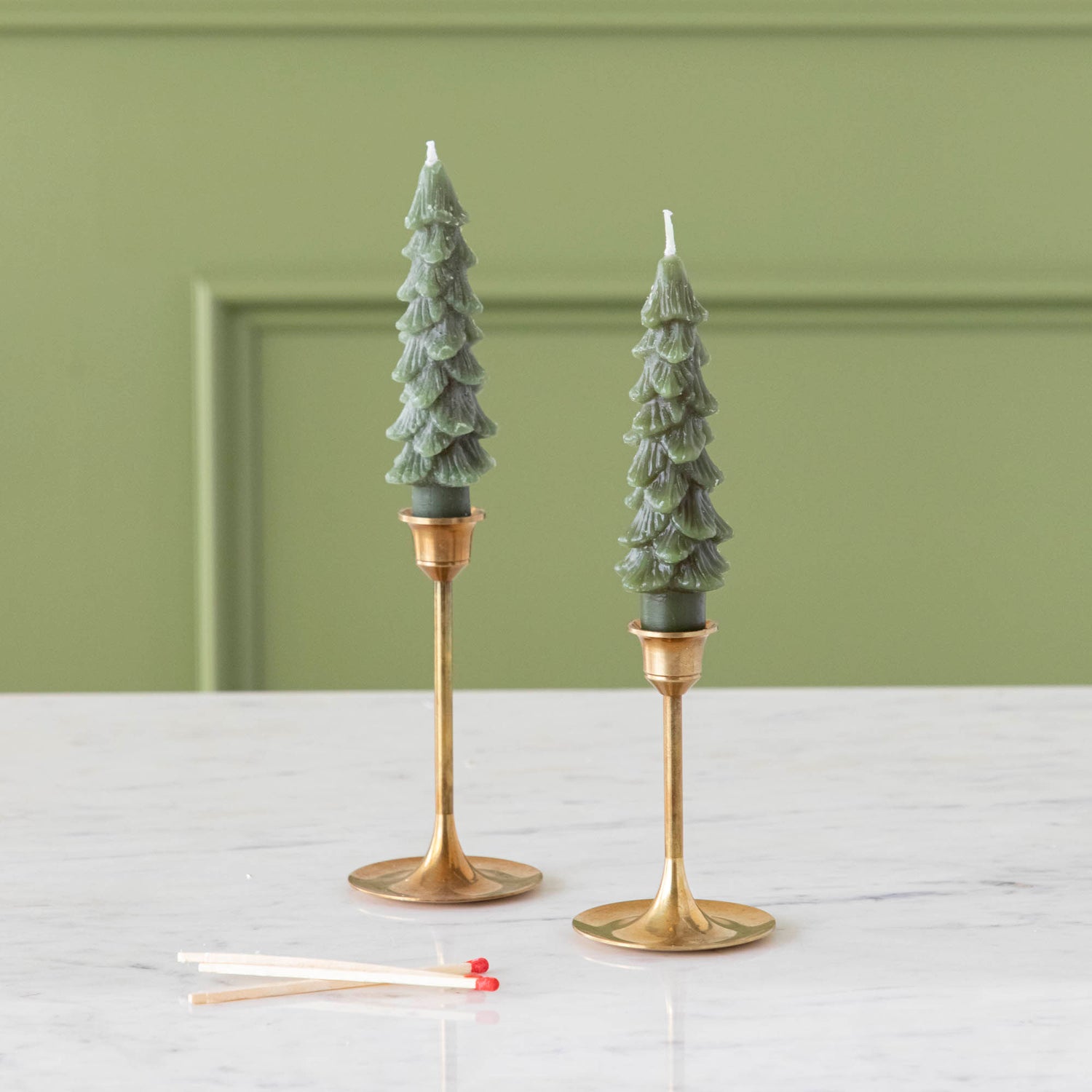 A set of two green tree-shaped taper candles placed on gold-colored candle holders.