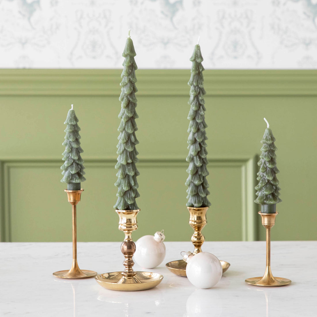 A set of two green tree-shaped taper candles placed on gold-colored candle holders, with a white ornamental ball beside them, set against a light green background.