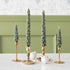 A set of two green tree-shaped taper candles placed on gold-colored candle holders, with a white ornamental ball beside them, set against a light green background.