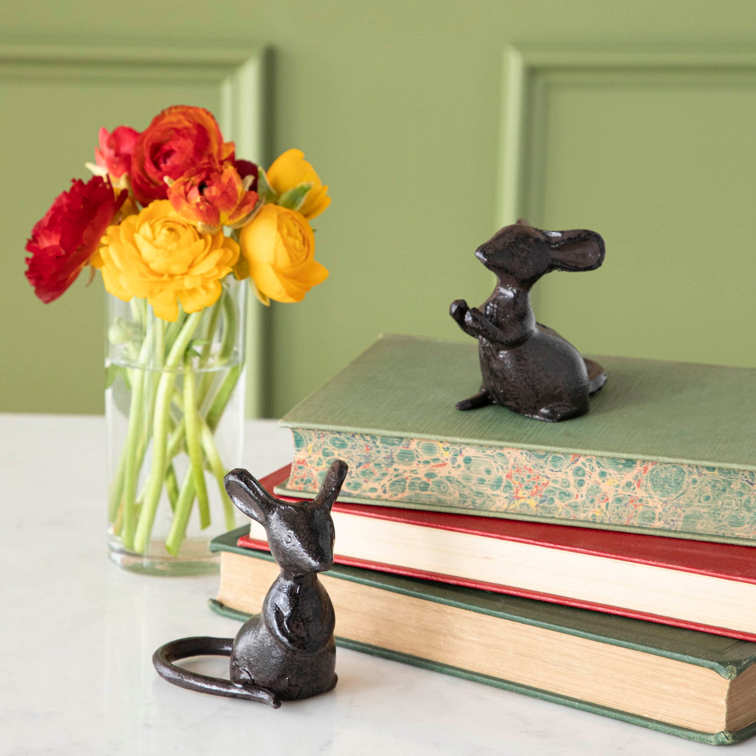 Two cast iron mice, one on top of a stack of books and one in front, next to a vase of flowers.