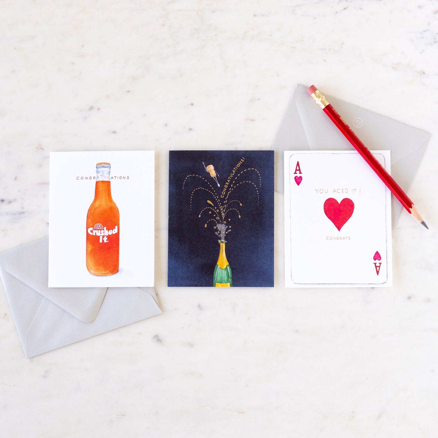 Three congratulatory cards in a row with two envelopes and a pencil, on a marble table.
