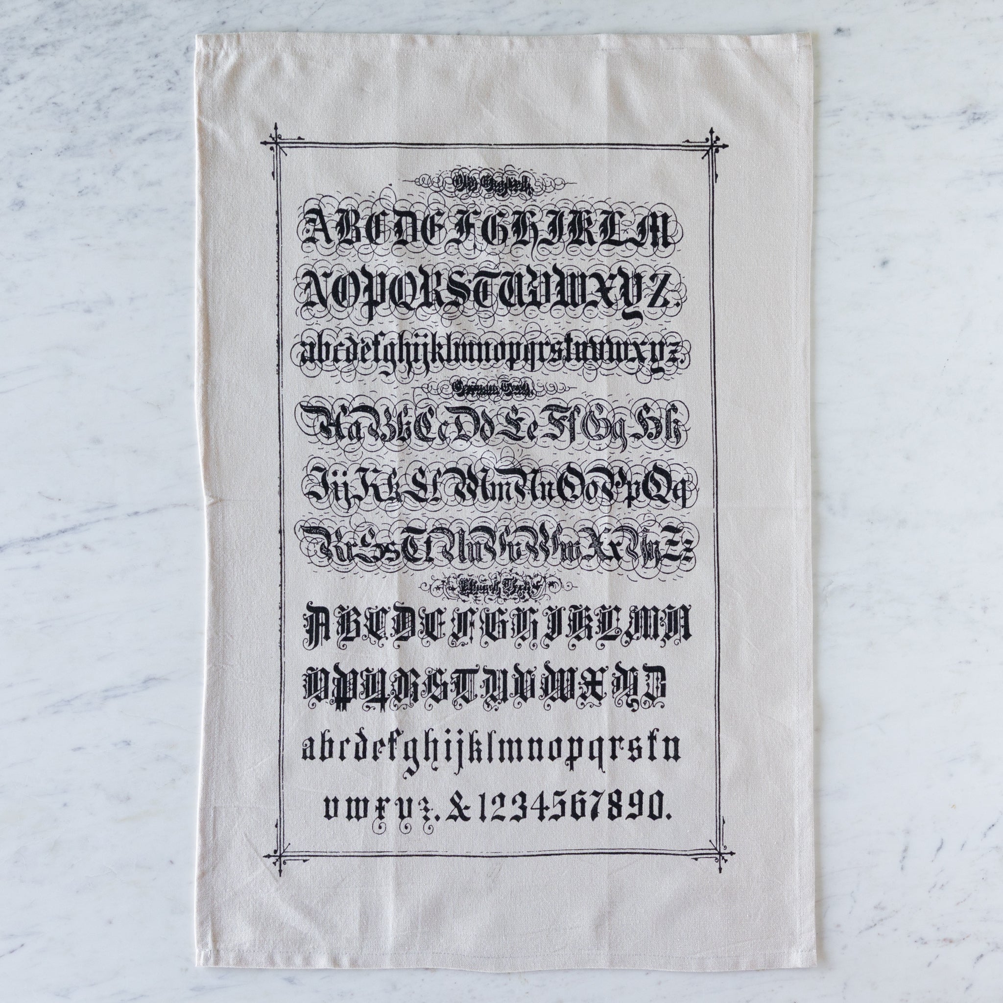 Old English Tea Towel featuring the alphabet and numbers in old english font on a marble table.