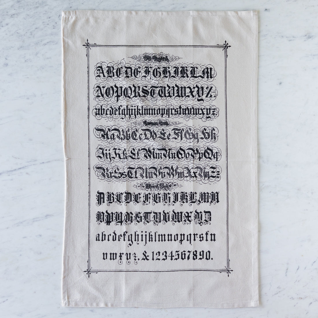 Old English Tea Towel featuring the alphabet and numbers in old english font on a marble table.