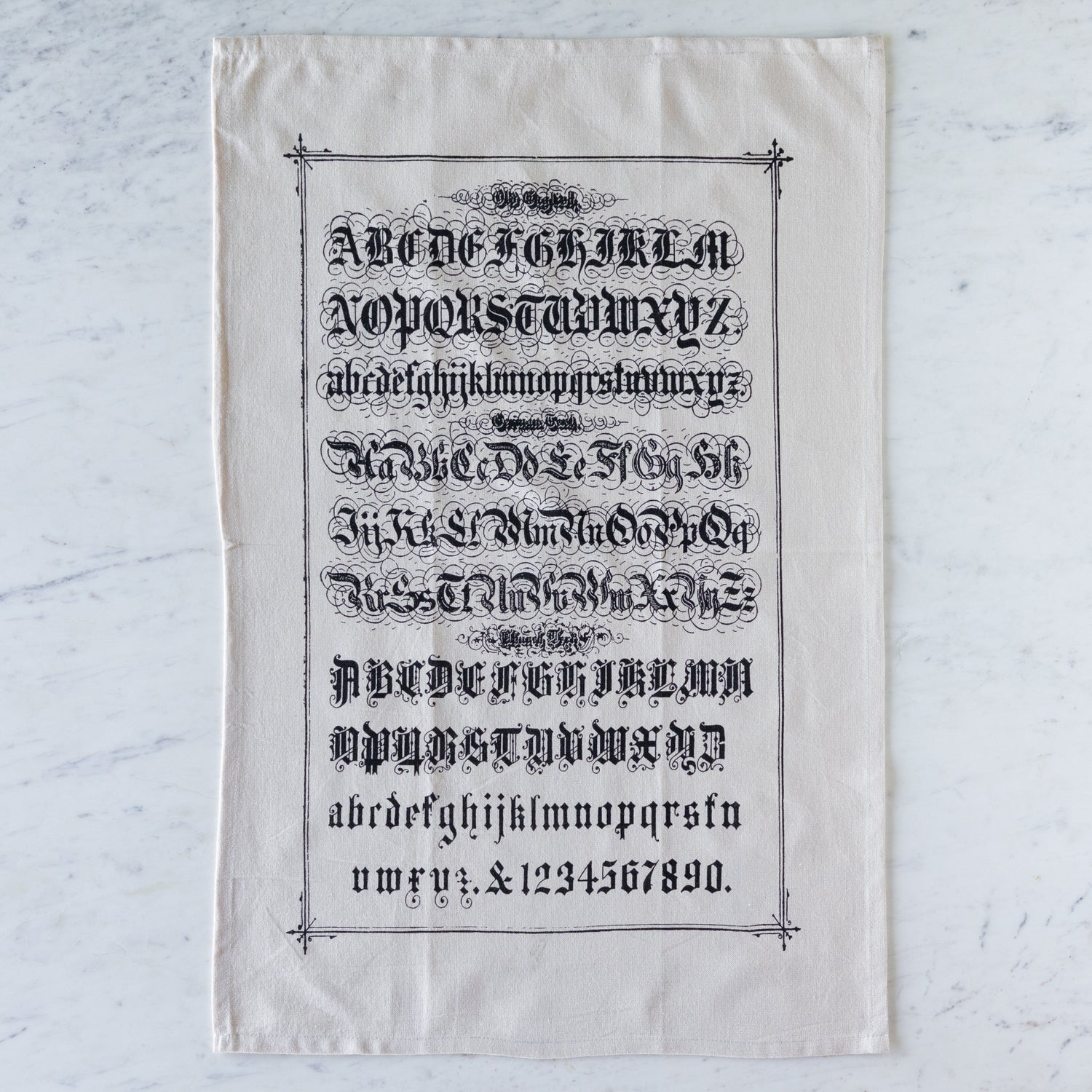 Old English Tea Towel featuring the alphabet and numbers in old english font on a marble table.