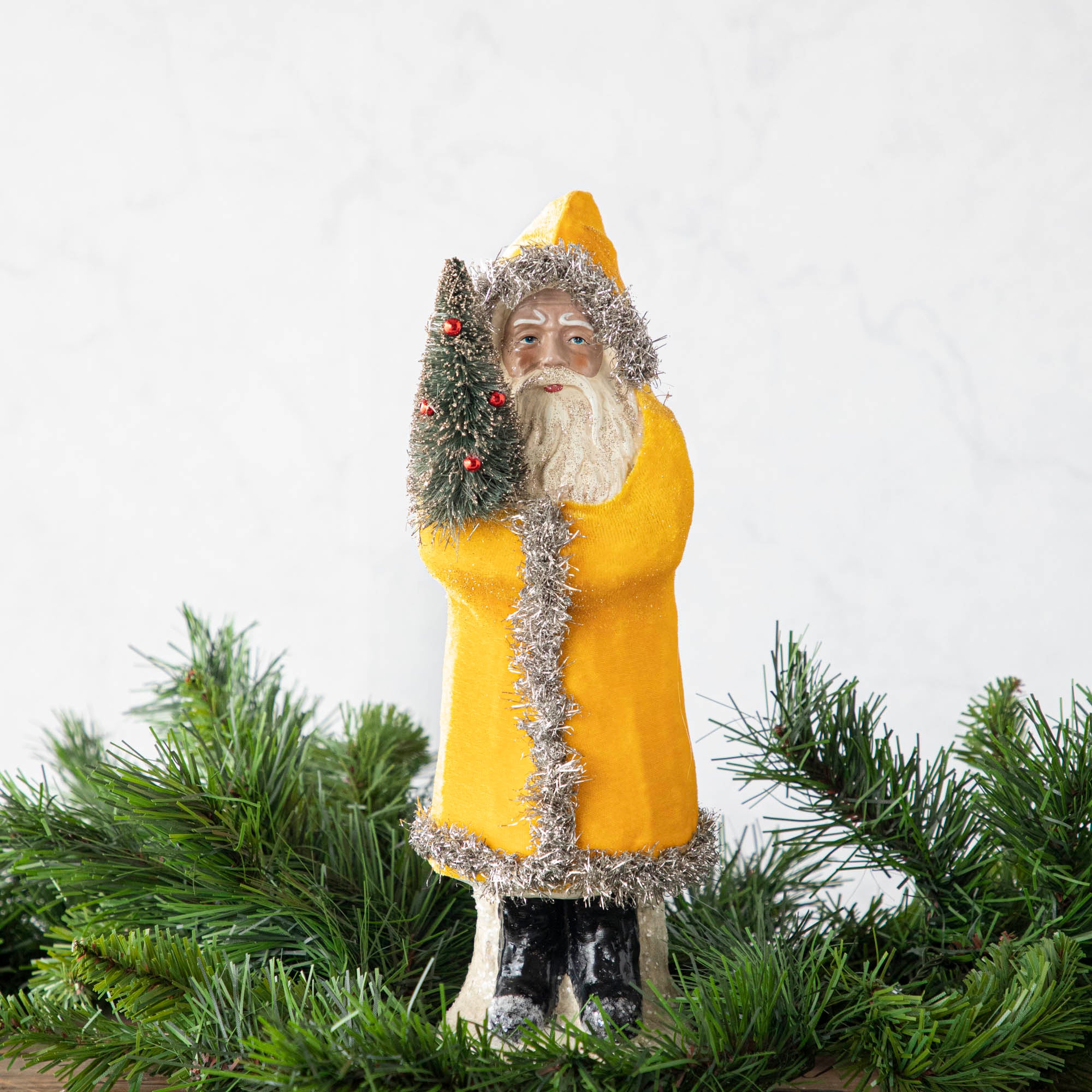 A 12" Velvet Belsnickle figurine with a yellow velvet robe adorned with tinsel trimming and glitter, holding a tree.