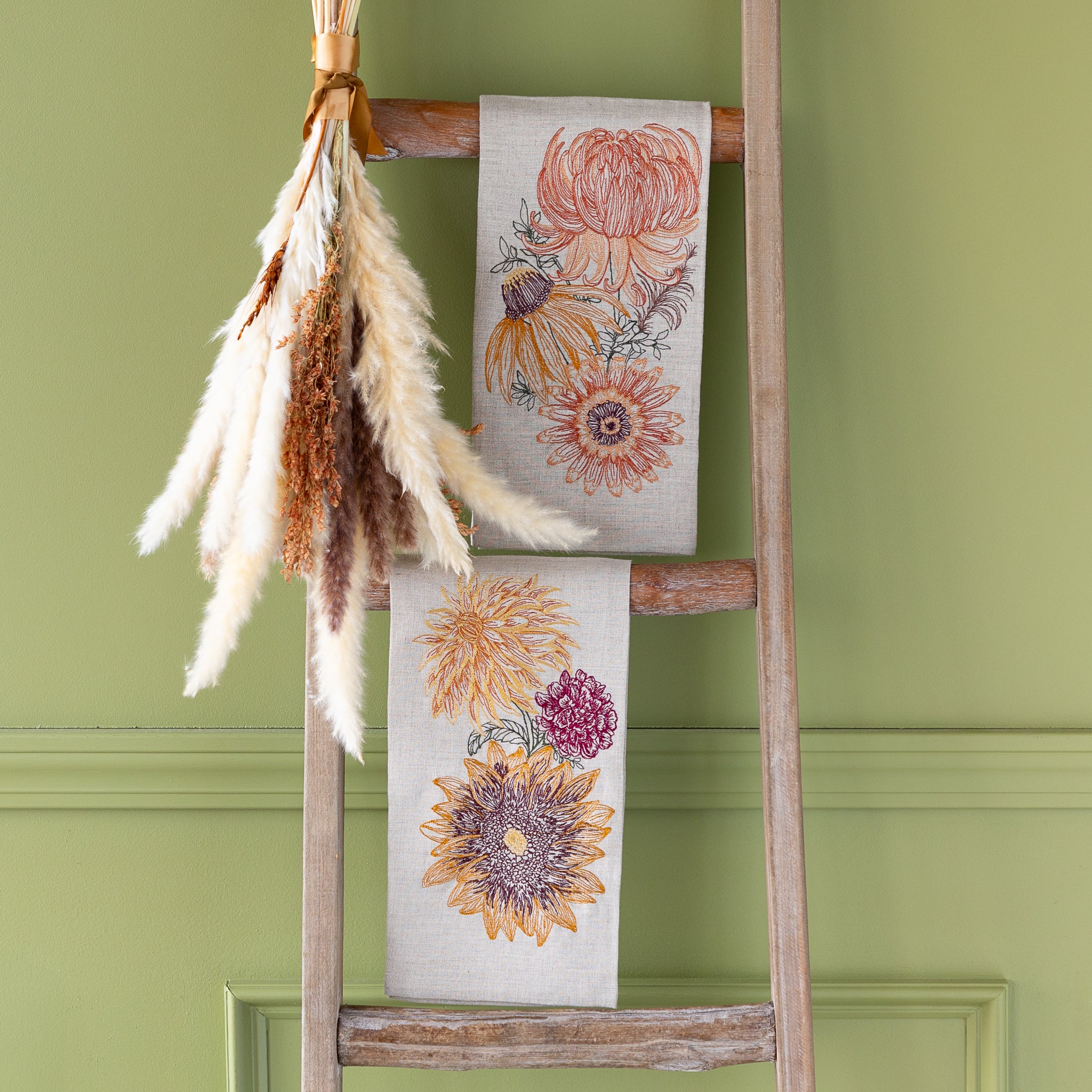 Autumn Sunflower Tea Towel and Autumn Mums Tea Towel hung on a wooden ladder against a green wall.