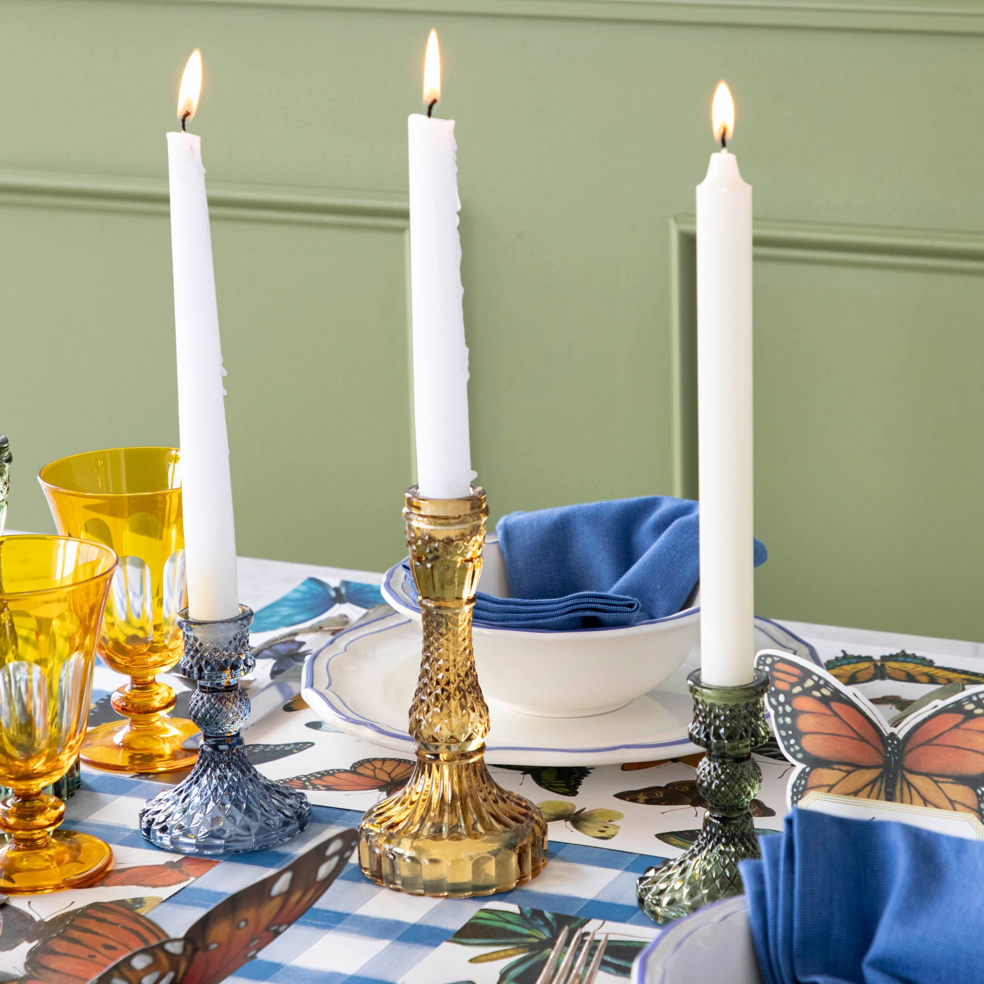 Three glass candle holders containing white lit candles on a set table.