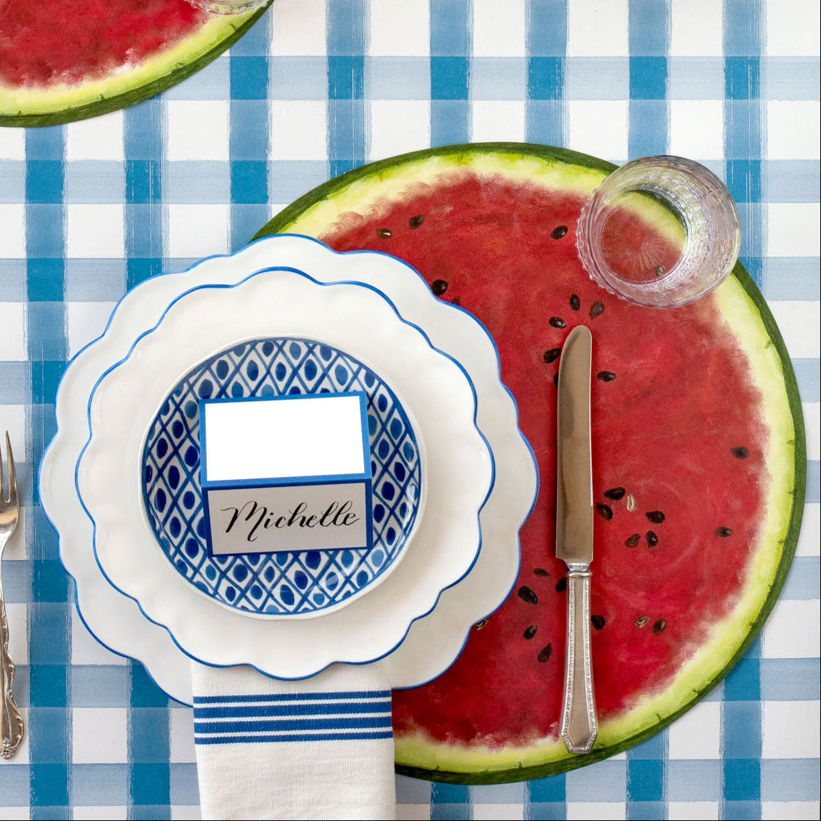 A summer place setting featuring the Die-cut Watermelon Placemat under blue scalloped plates, a knife, empty glass, napkin and Blue Frame Place Card atop, on the Blue Painted Check runner.