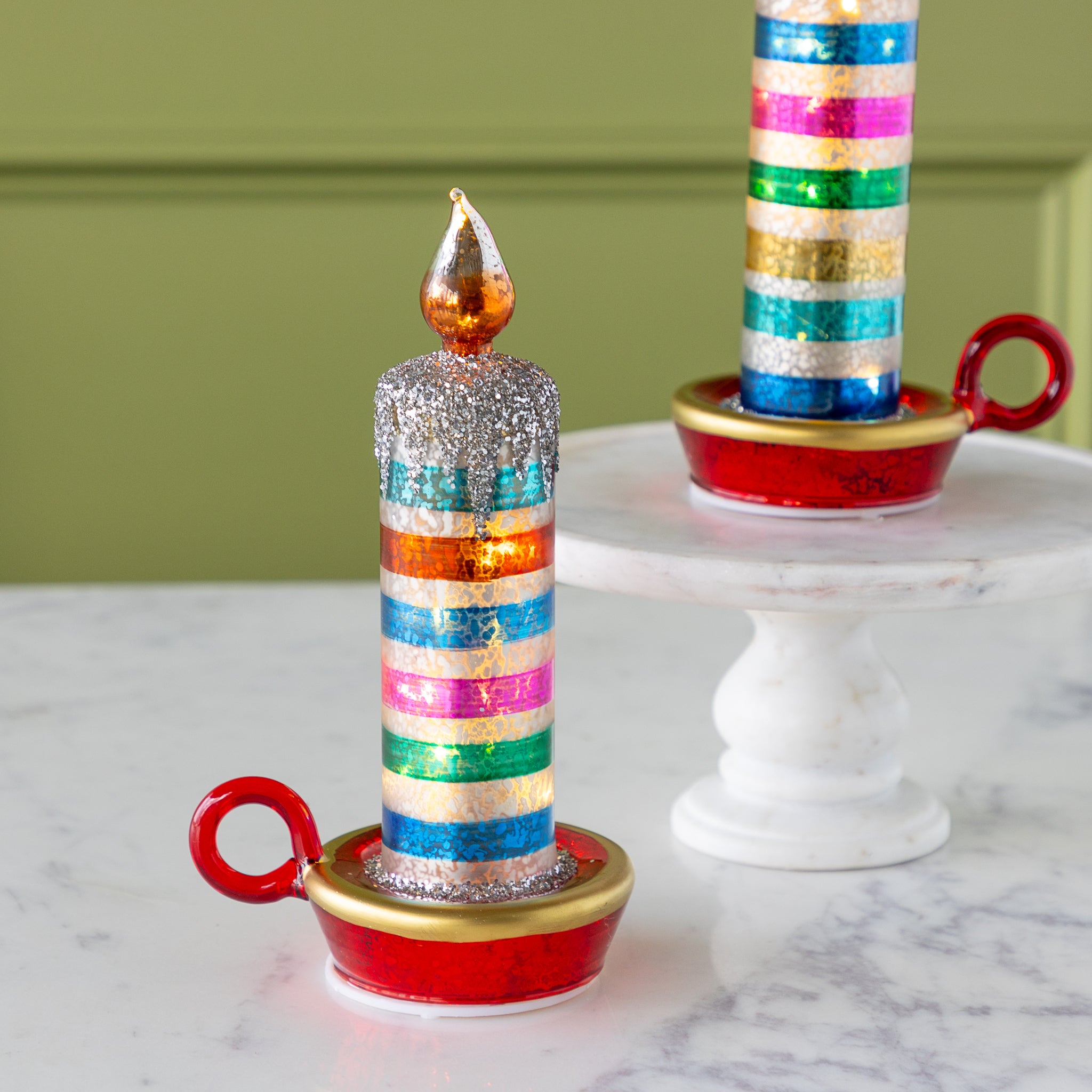 Two decorative retro stripe candles with glittery tops and gold-painted flames sit on red and gold holders; one is on a marble pedestal, the other on a marble surface.