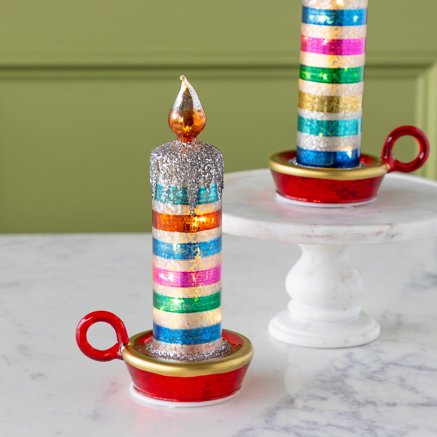 Two decorative retro stripe candles with glittery tops and gold-painted flames sit on red and gold holders; one is on a marble pedestal, the other on a marble surface.
