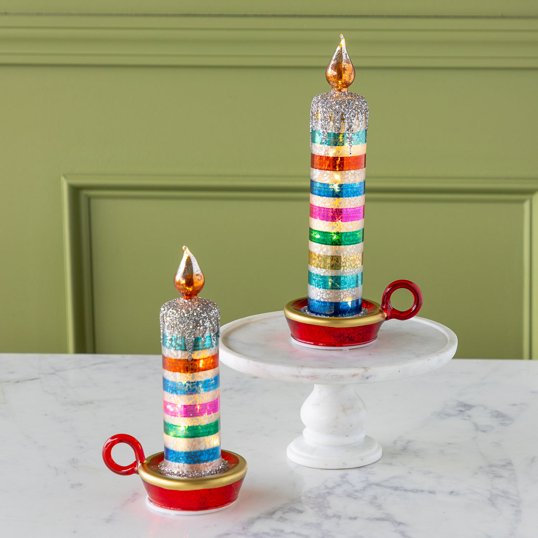Two decorative retro stripe candles with glittery tops and gold-painted flames sit on red and gold holders; one is on a marble pedestal, the other on a marble surface.