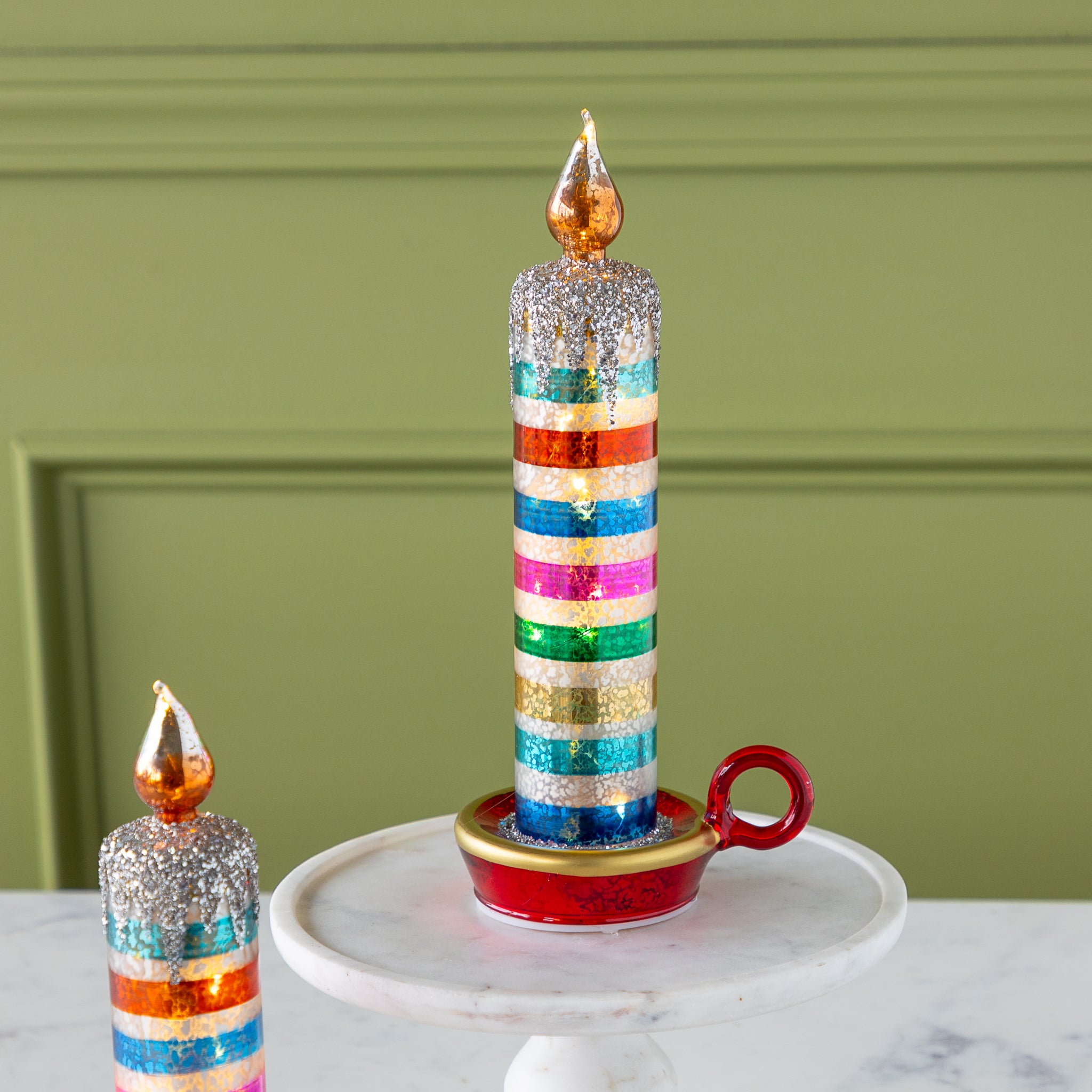 Two decorative retro stripe candles with glittery tops and gold-painted flames sit on red and gold holders; one is on a marble pedestal, the other on a marble surface.