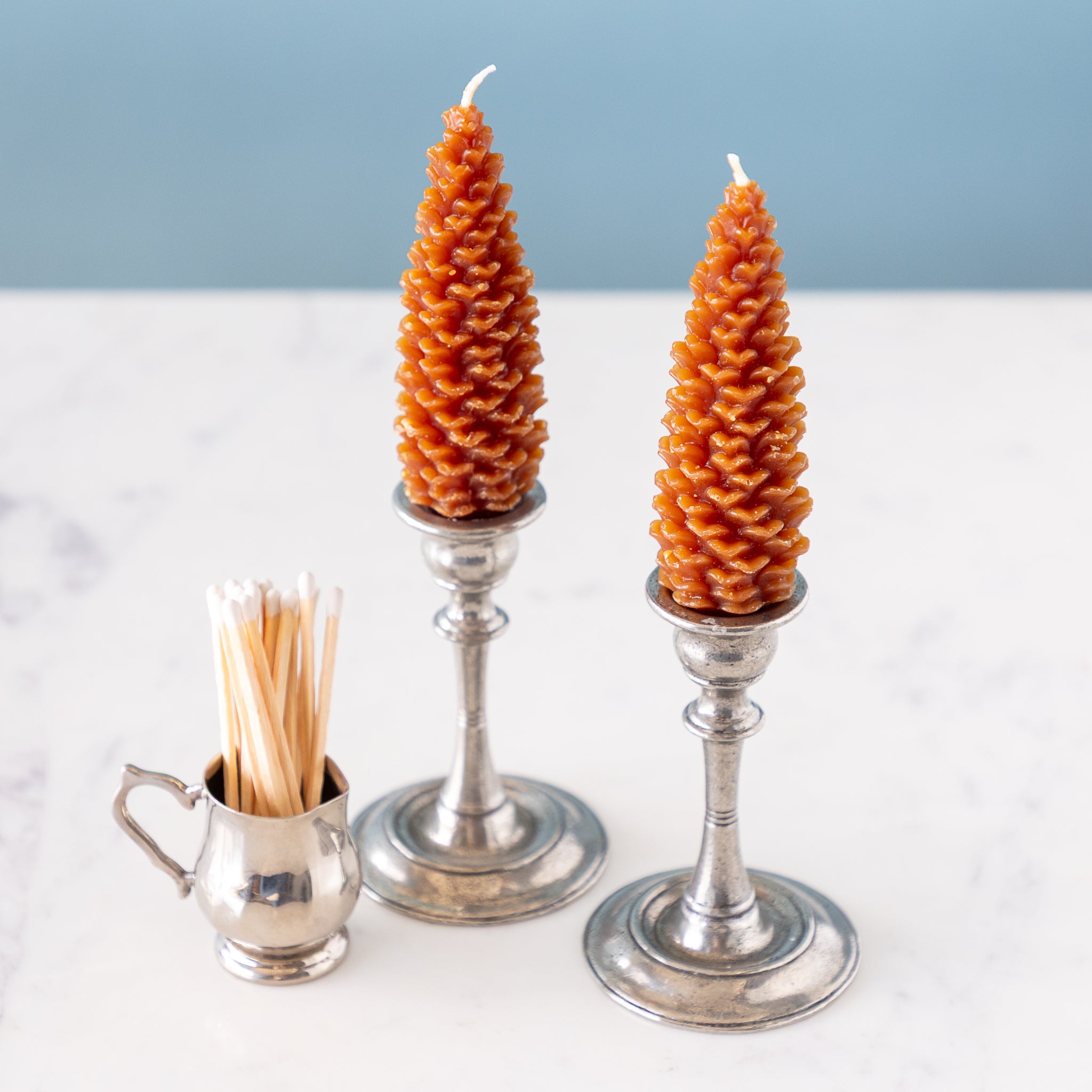 Two pinecone beeswax candles on silver candle holders with a small silver container of matches on a light background.