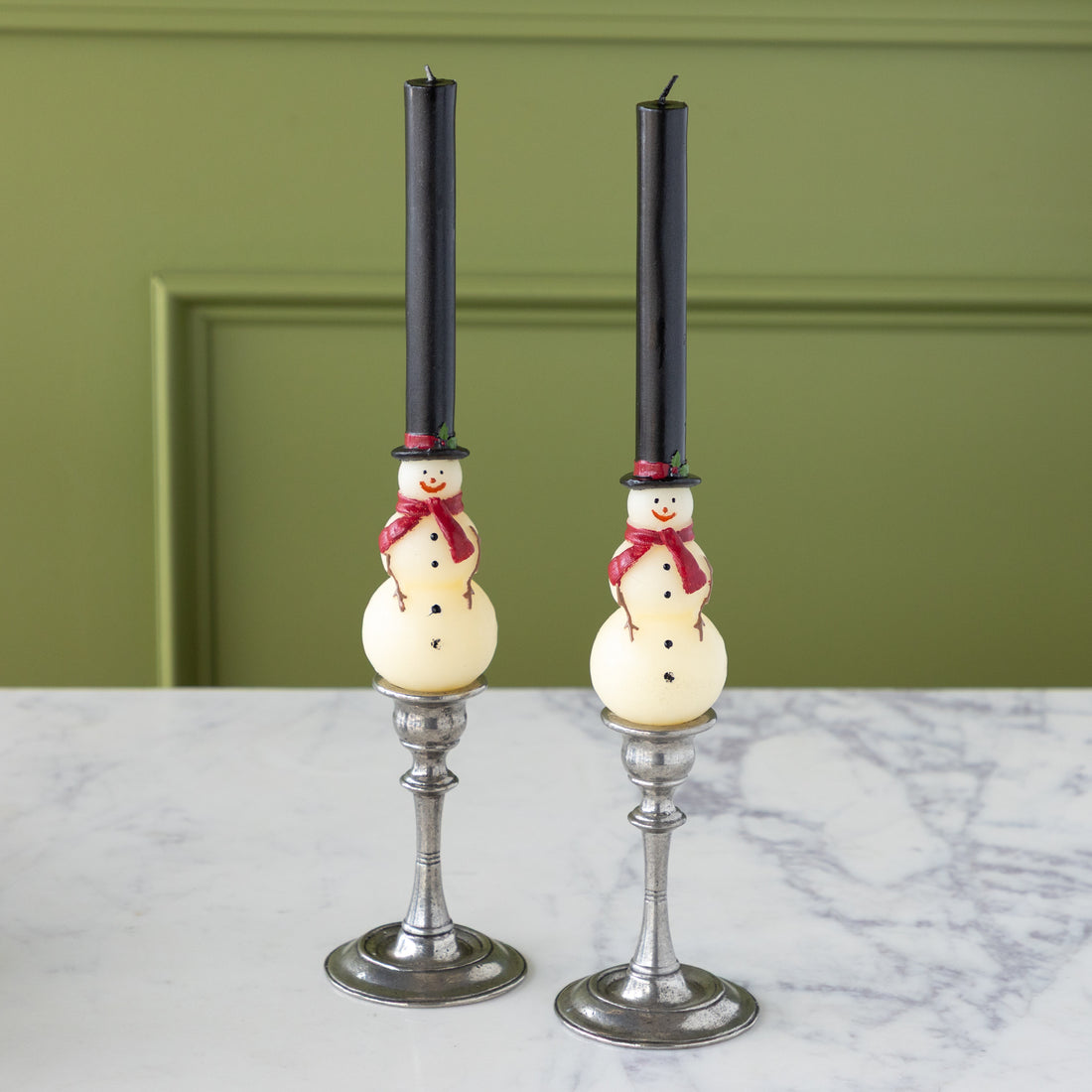 Two Snowman Taper Candles in vintage candlestick holders on a marble table against a green wall.