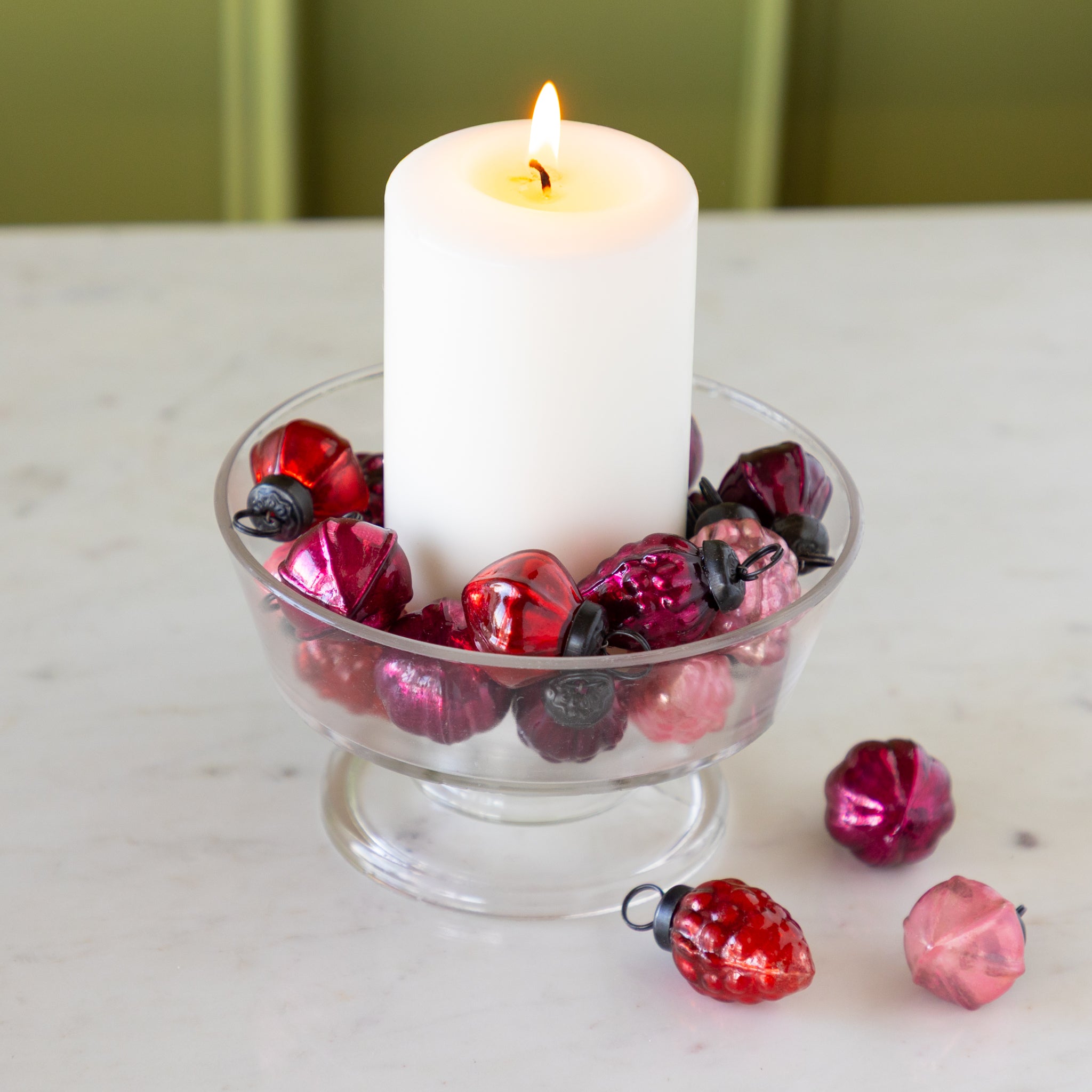 White candle in a glass holder with Multi Color Mercury Glass Ornaments on a marble surface.