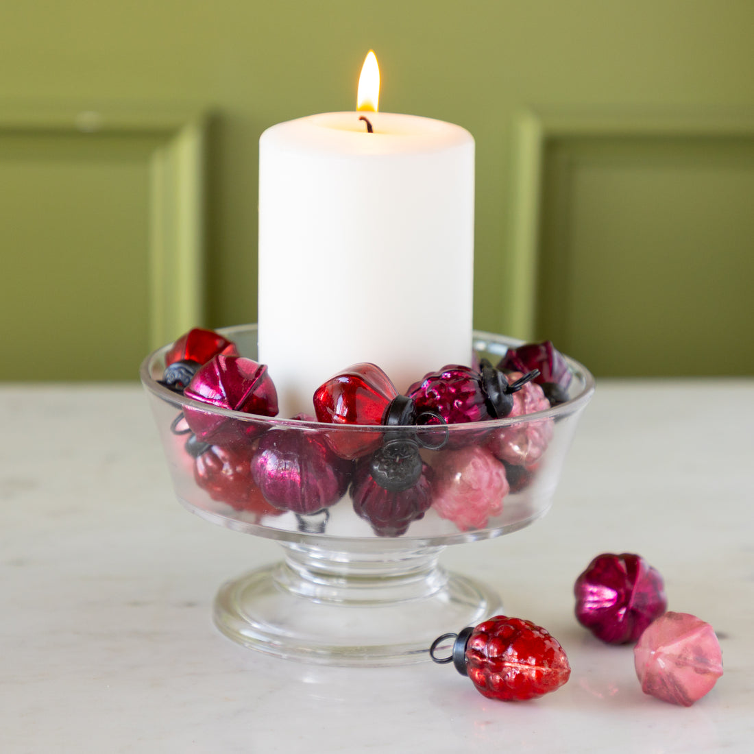 White candle in a glass holder with Multi Color Mercury Glass Ornaments on a marble surface.