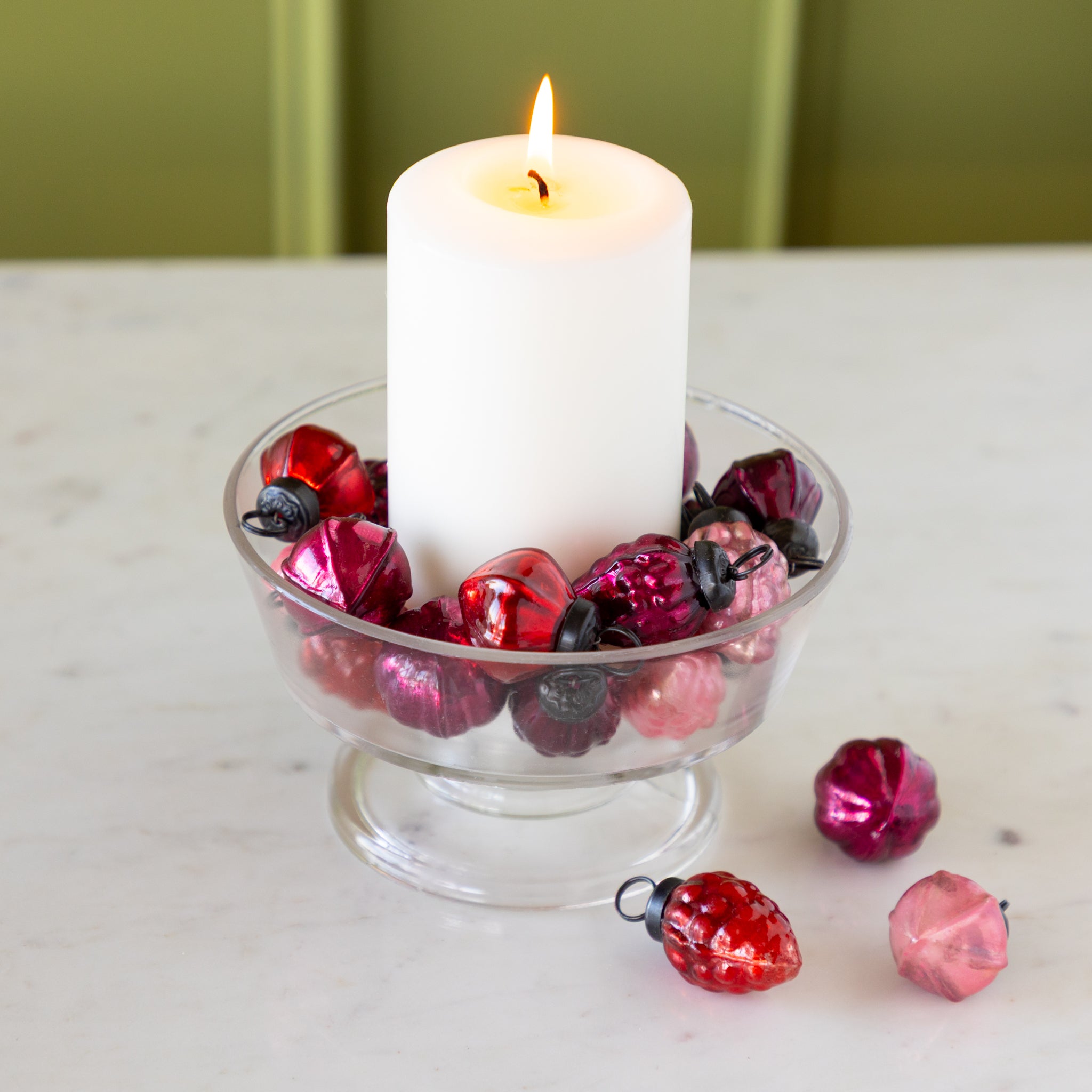 White candle in a glass holder with Multi Color Mercury Glass Ornaments on a marble surface.