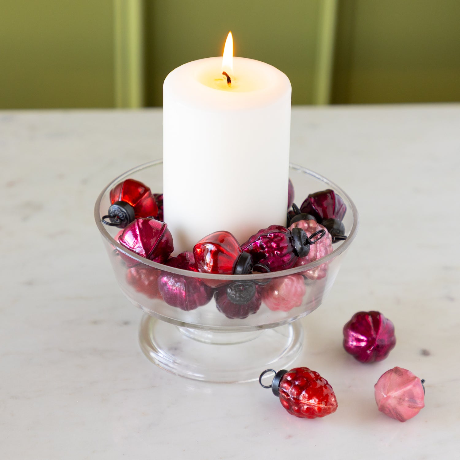 White candle in a glass holder with Multi Color Mercury Glass Ornaments on a marble surface.