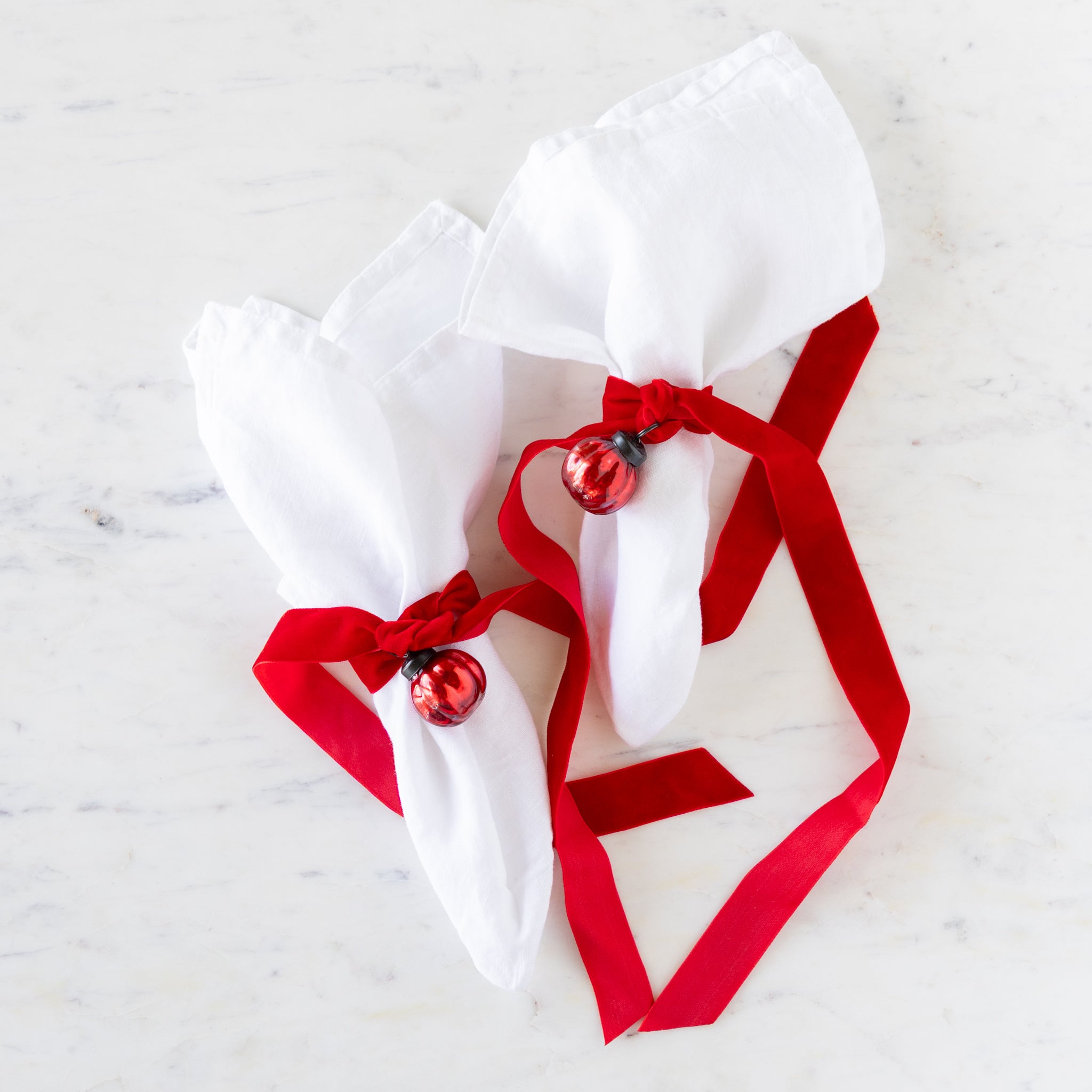 Two white linen napkins tied with red velvet ribbon and a red mercury glass ornament on a marble table.