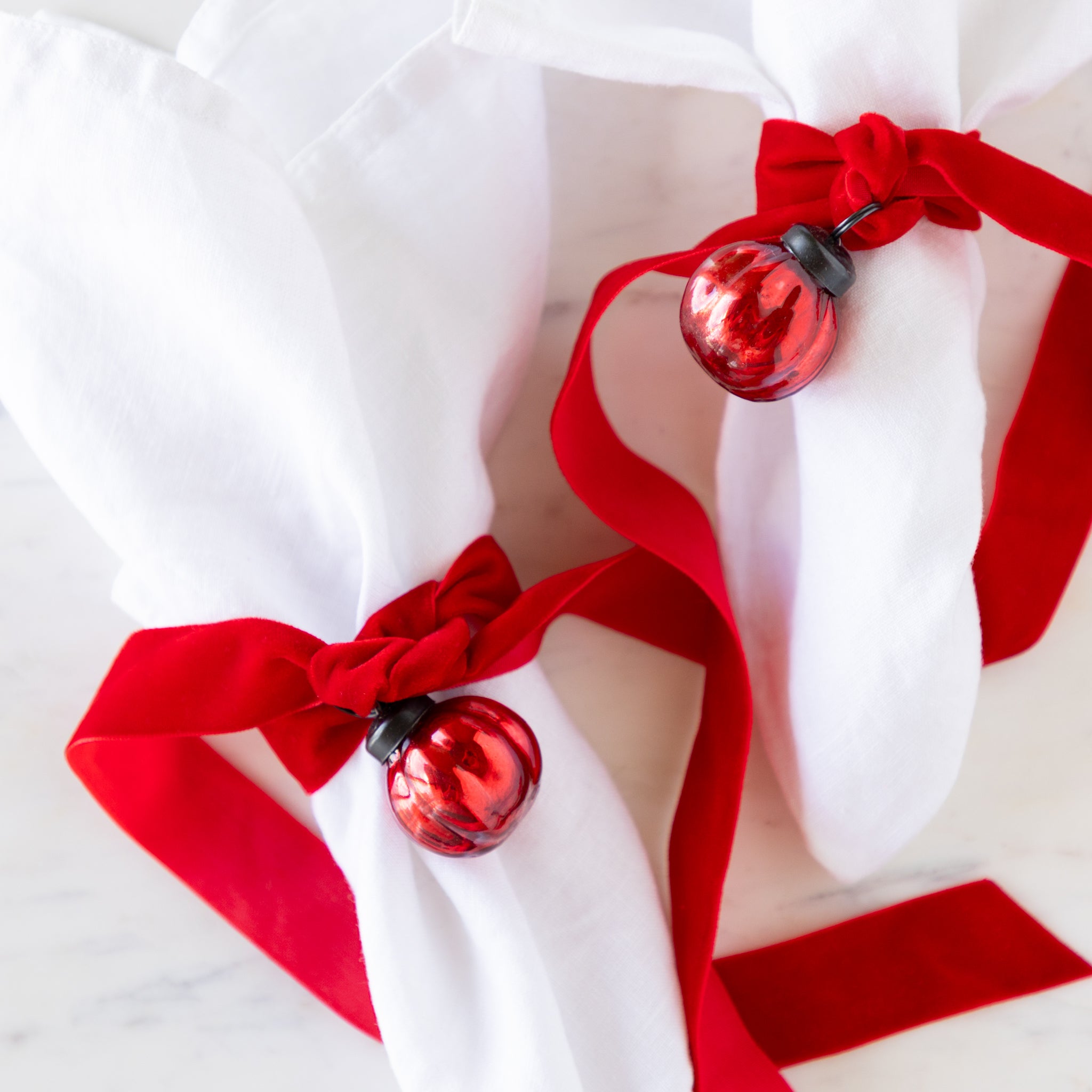 Two white linen napkins tied with red velvet ribbon and a red mercury glass ornament on a marble table.