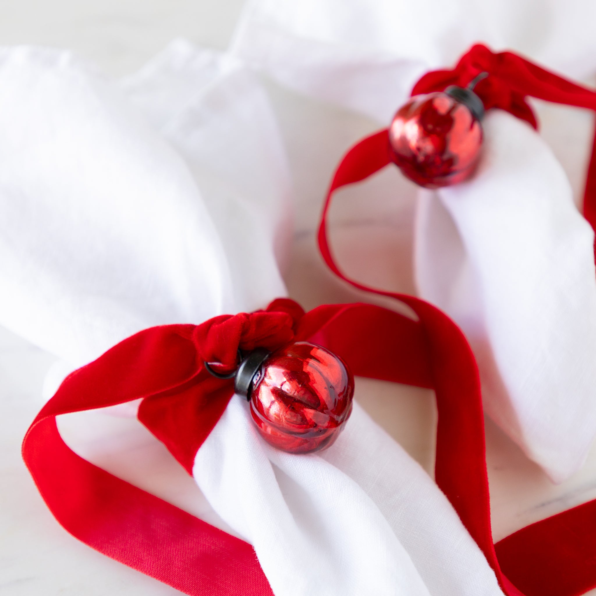 Two white linen napkins tied with red velvet ribbon and a red mercury glass ornament on a marble table.