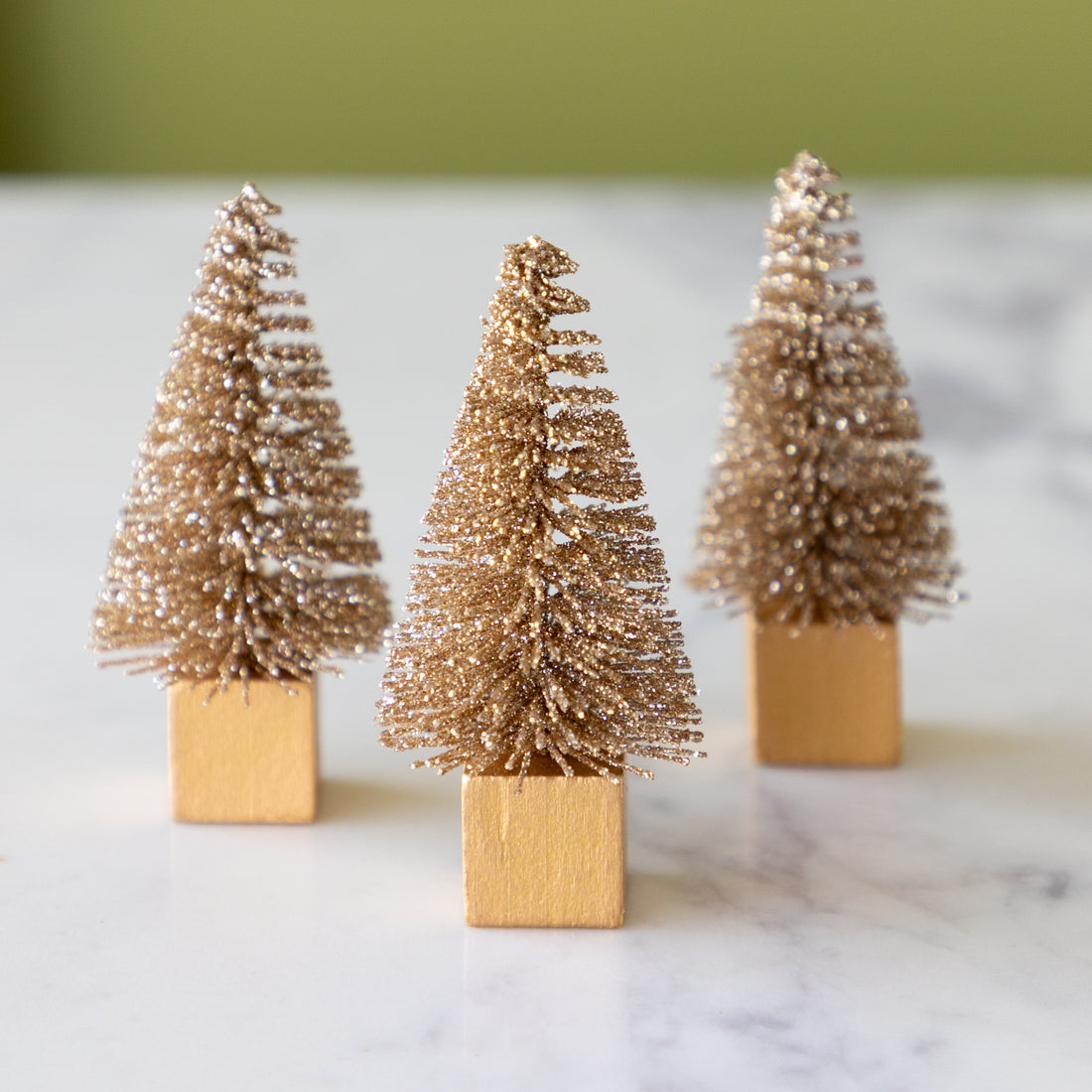 Three miniature gold bottle brush trees on wooden stands against a white background.