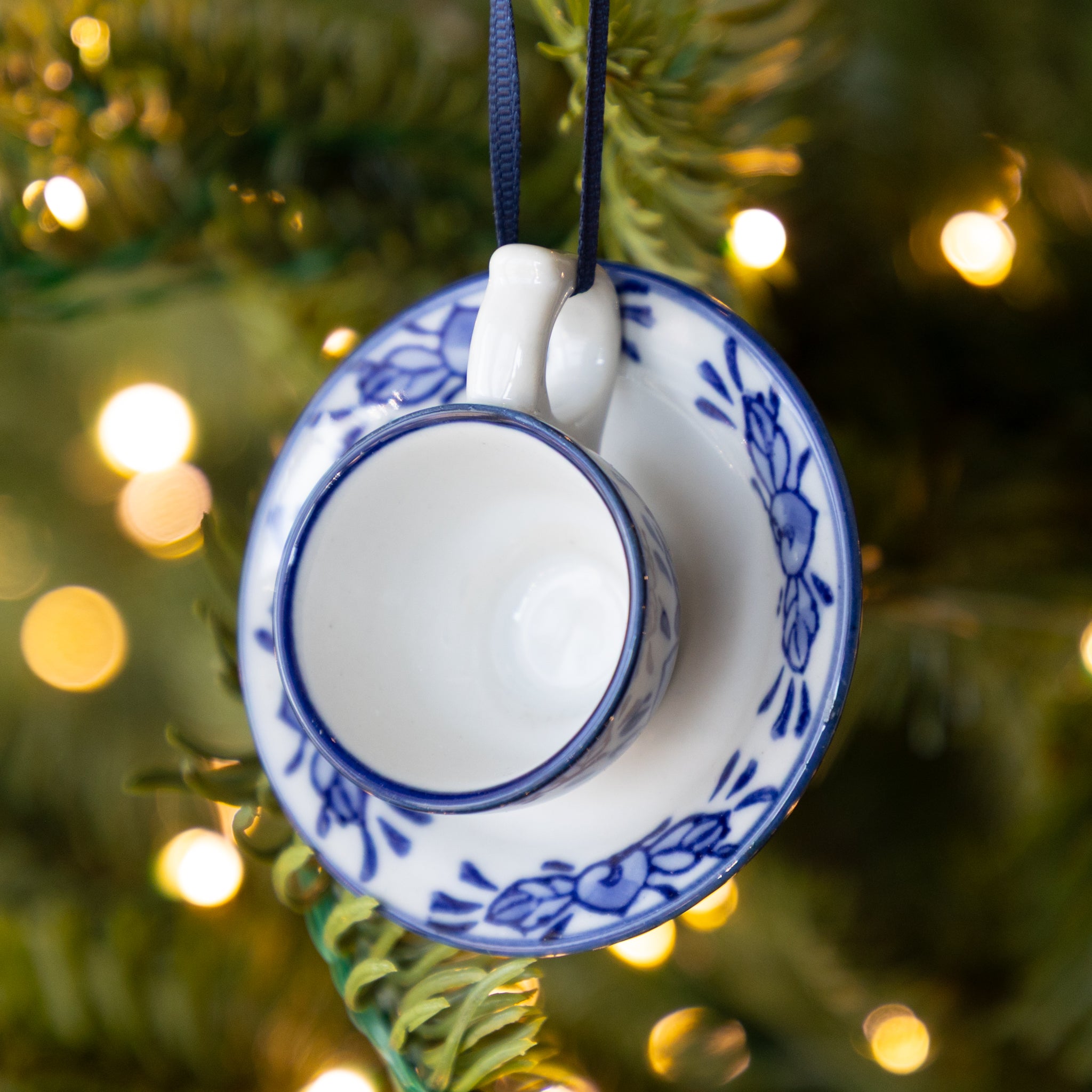 Decorative teacup ornament hanging on a Christmas tree with blurred lights in the background.