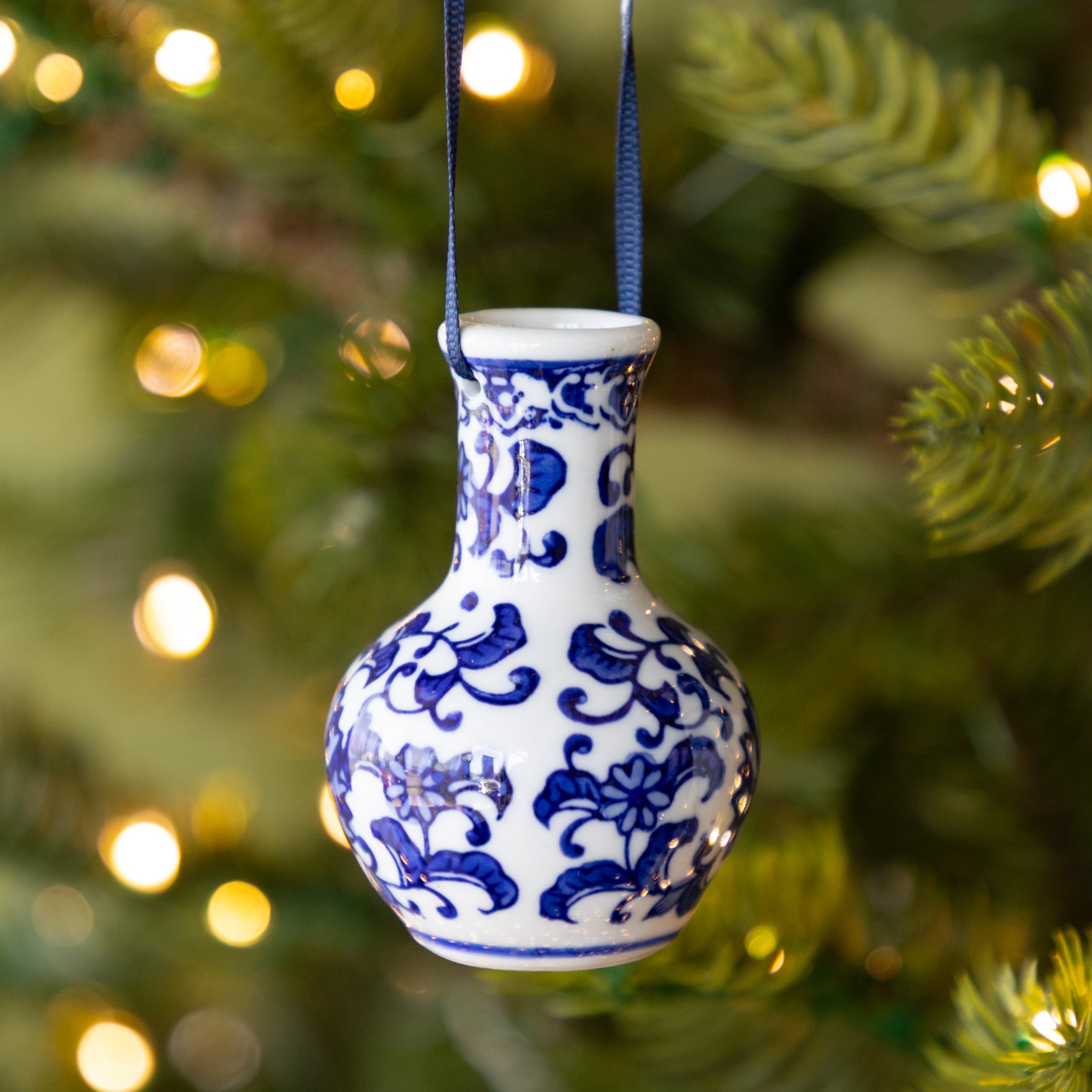Blue and white ceramic vase ornament hanging on a Christmas tree with blurred lights in the background.