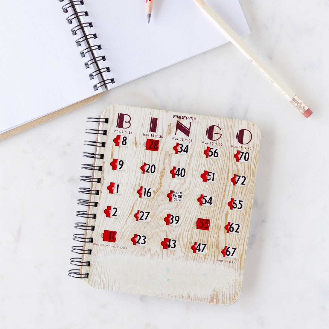 Bingo Notebook featuring a classic Bingo board cover with red markers to cover the numbers and a spiral bound, on a marble table next to an open notebook and pencil.